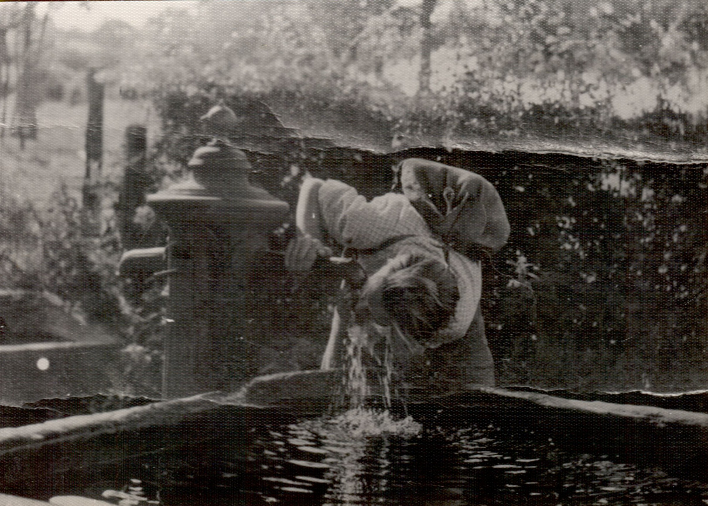 A young adult bends down to drink water from a fountain in an outdoor setting.