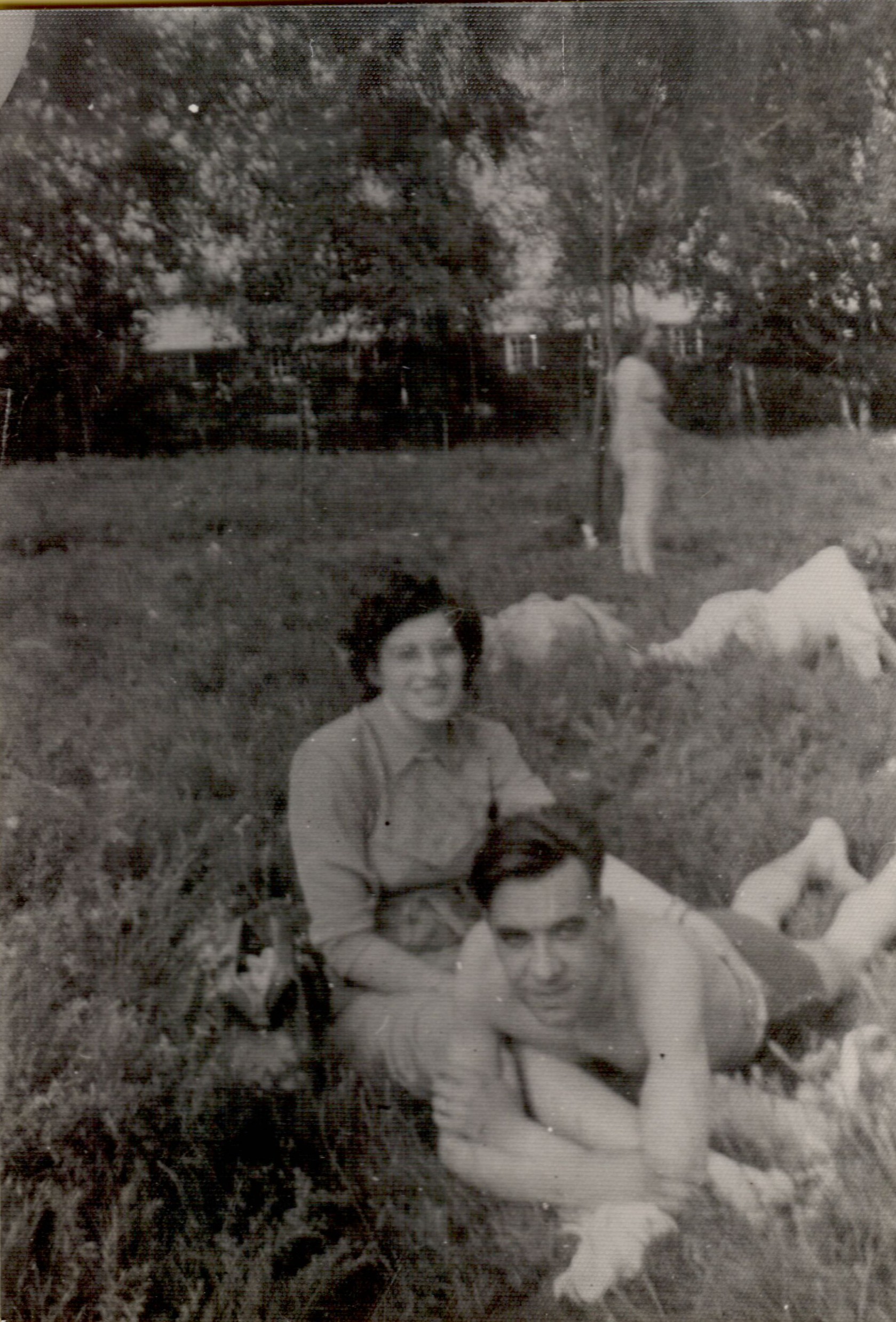 A woman sits on the grass, smiling, while a shirtless man lies in front of her, resting on his arms.