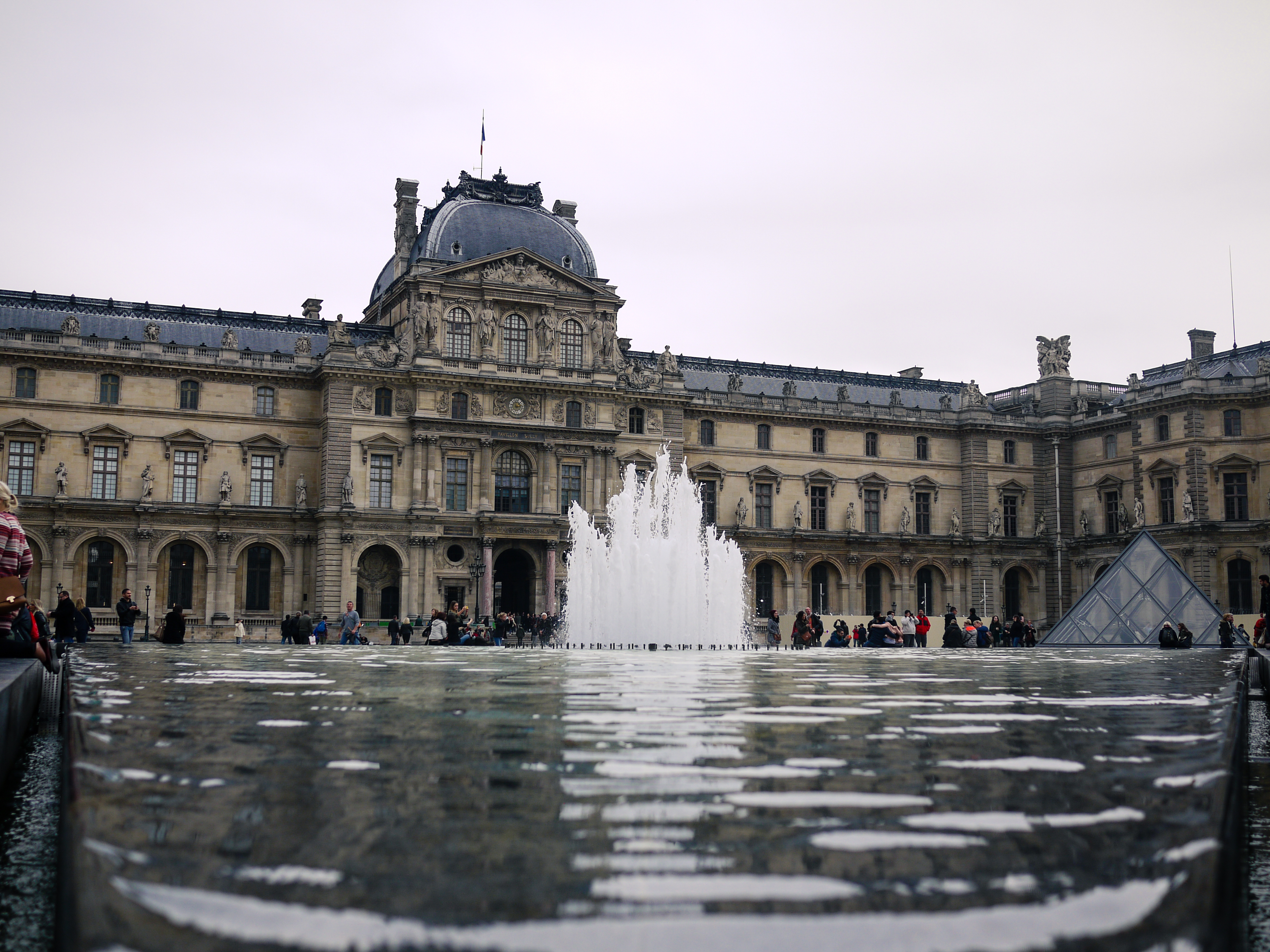 A large fountain sprays water in front of the Louvre Museum, with people walking nearby.