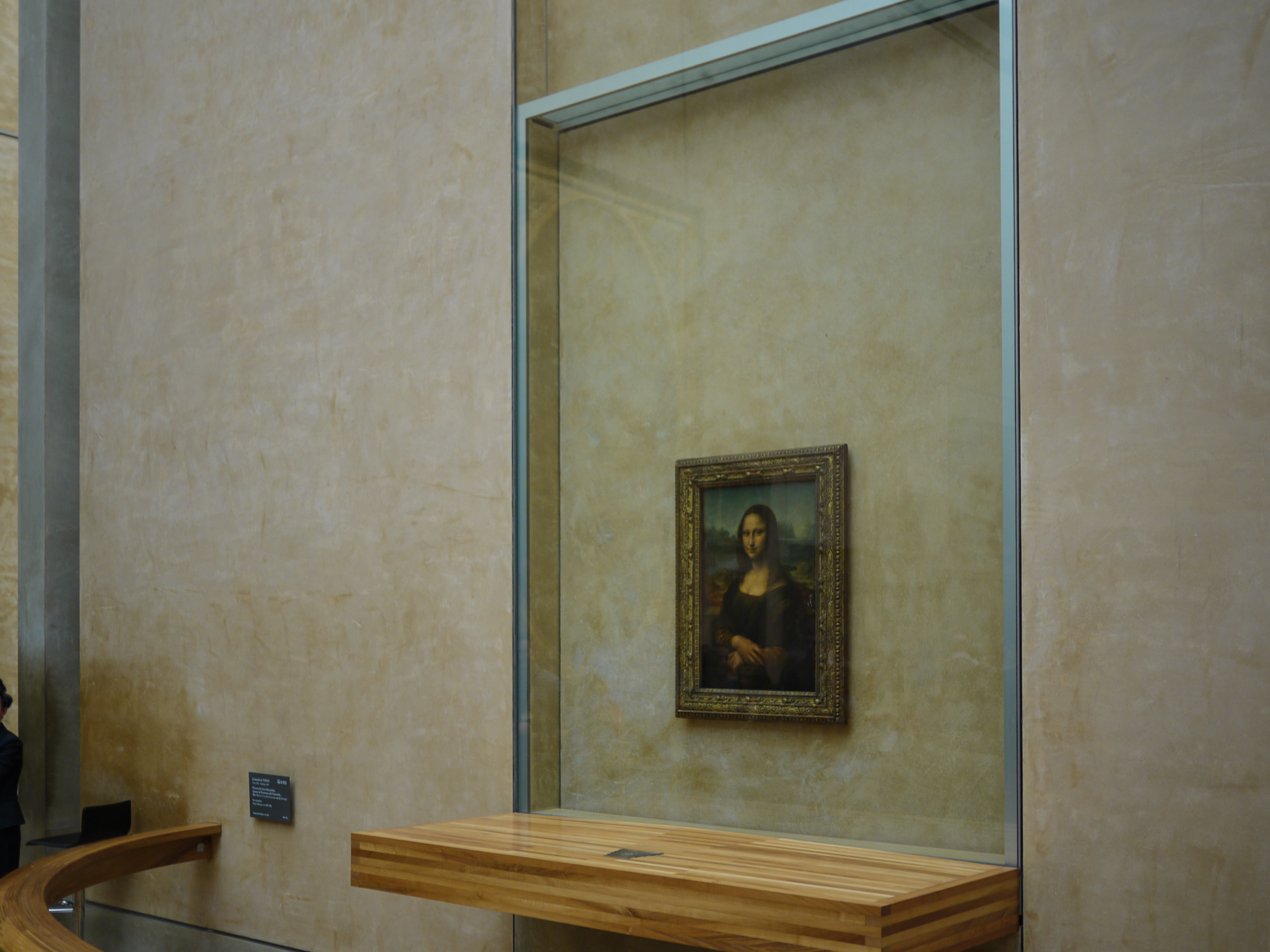 The Mona Lisa painting is displayed behind glass in a museum with a wooden barrier in front.
