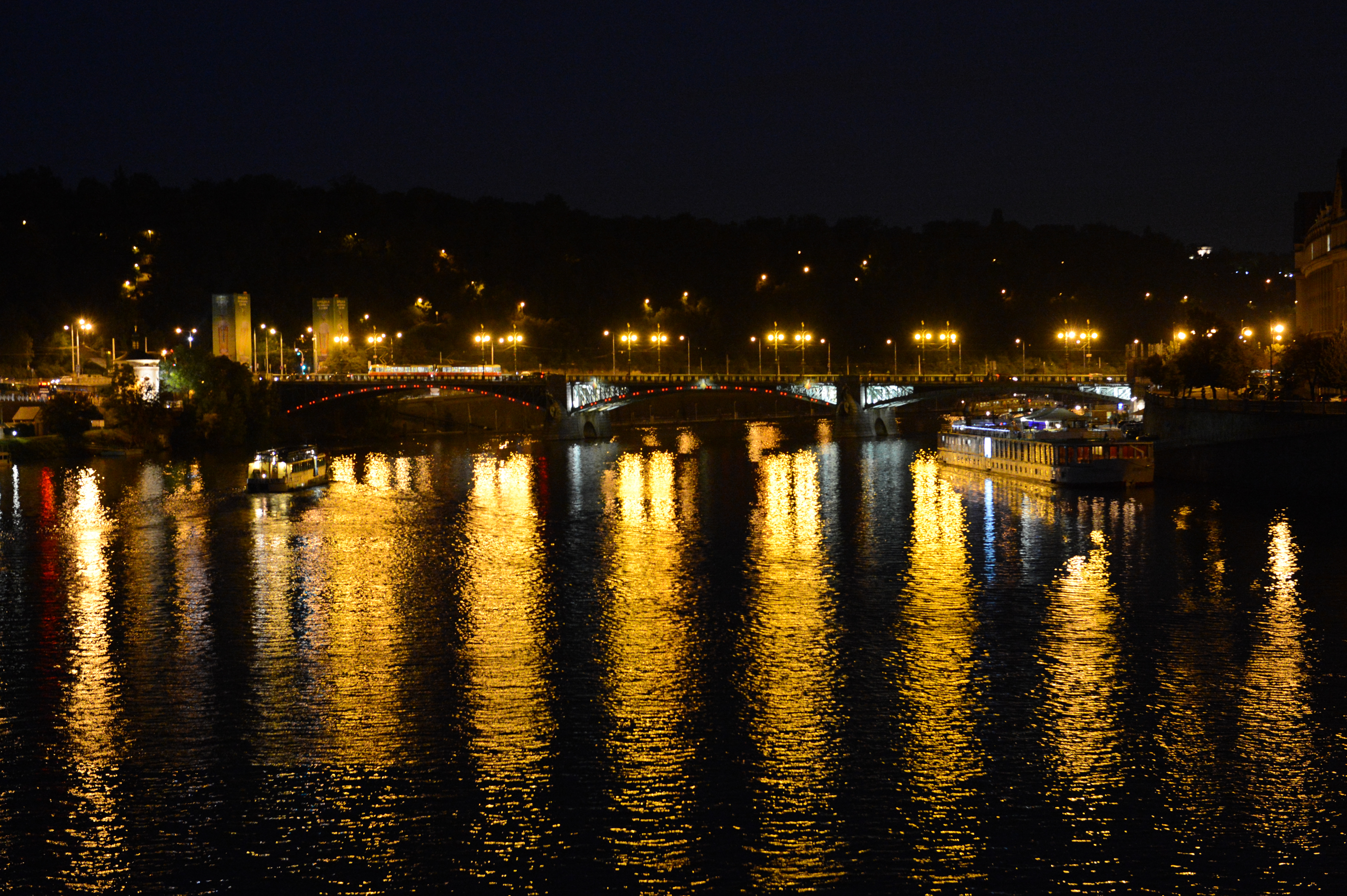 A bridge in Prague at night with streetlights reflecting on the river, and boats moving through the water.