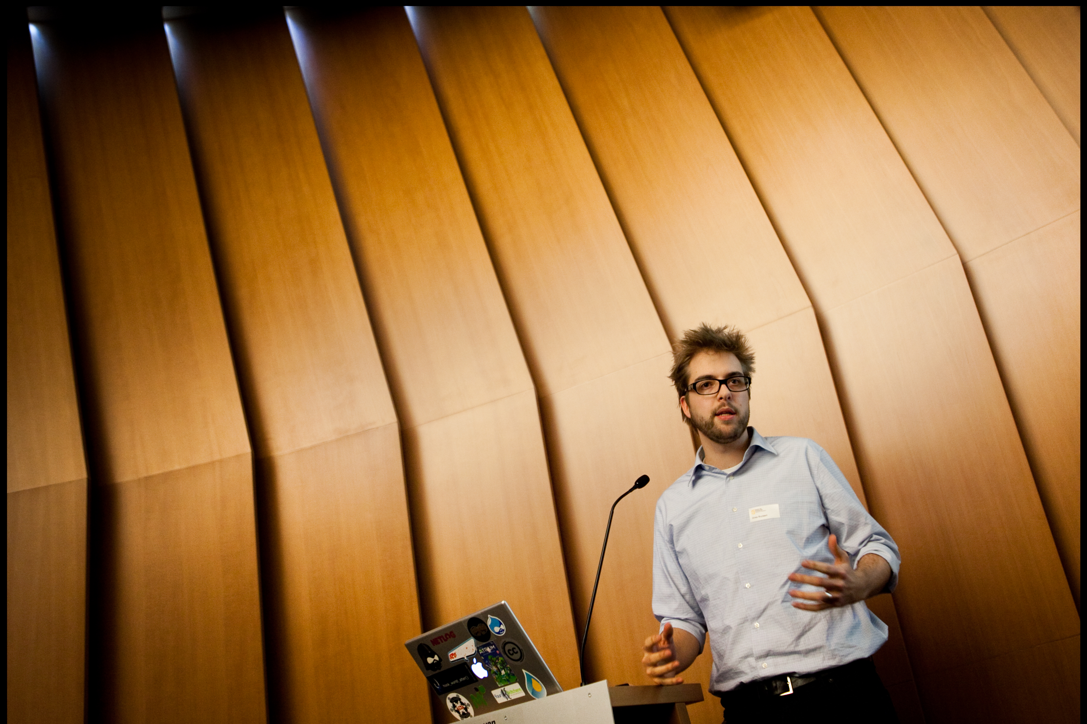 A man in a light blue shirt gives a presentation at a podium with a laptop and microphone.