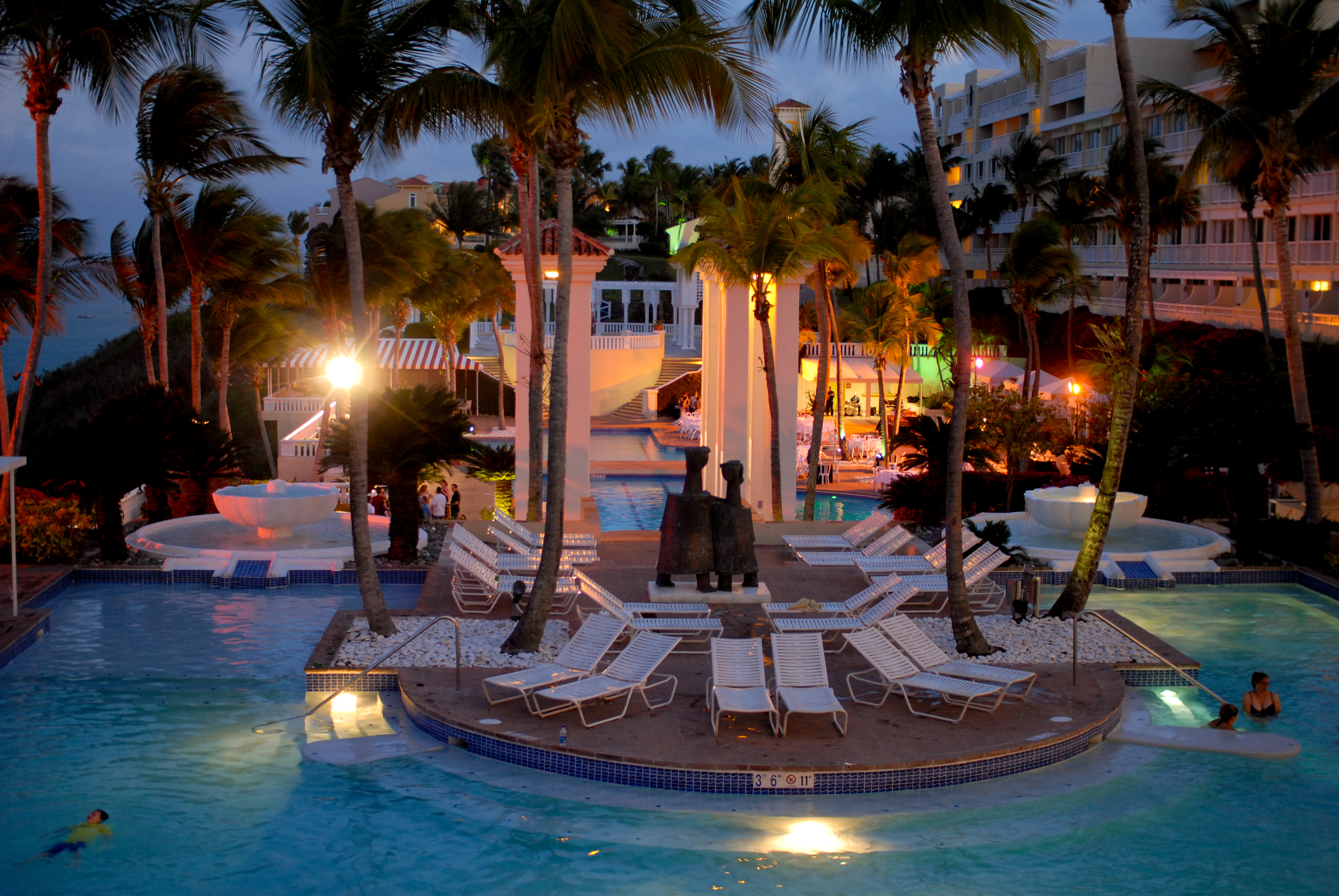 A resort pool area at night with lounge chairs, fountains, and people swimming and relaxing in the water.