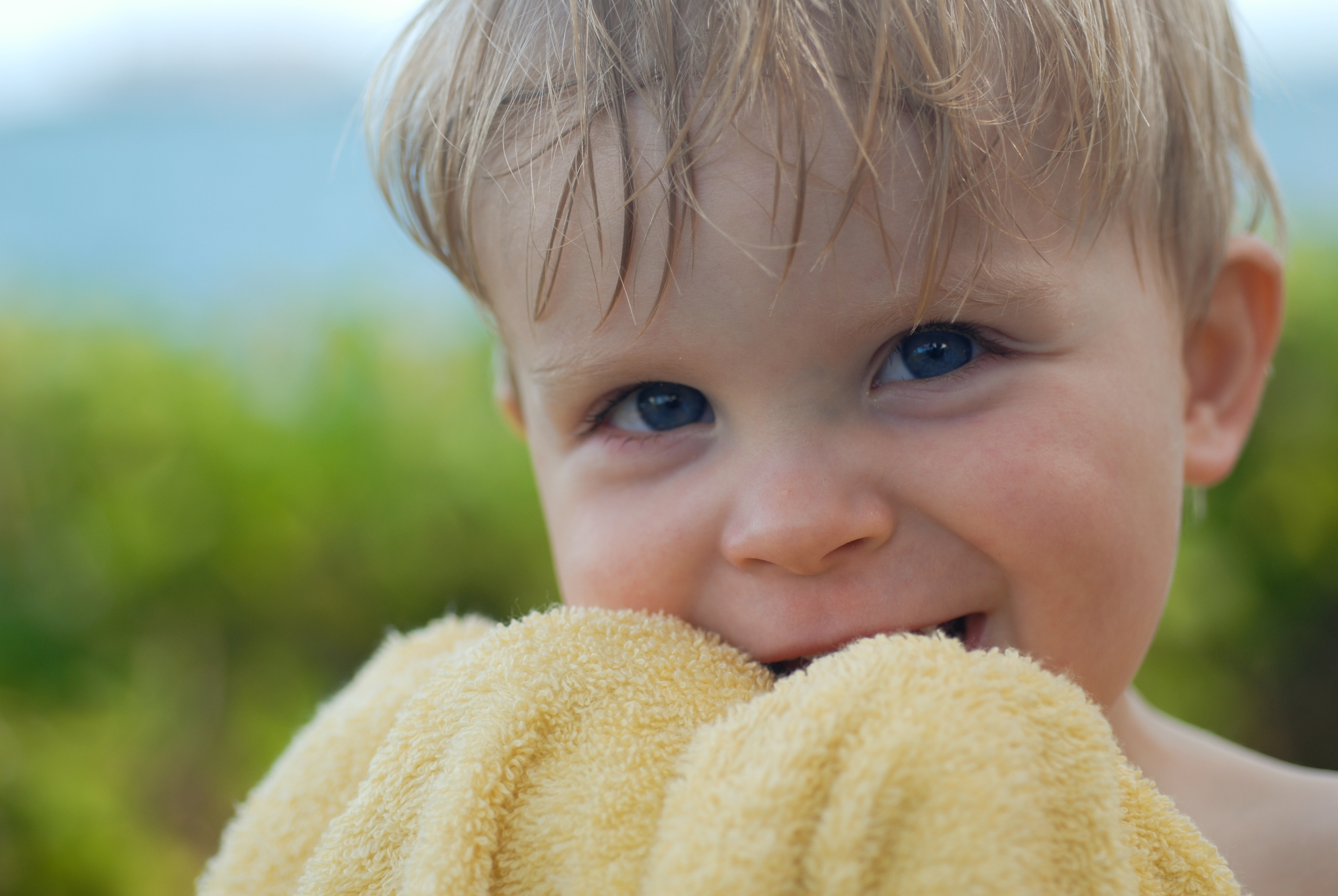 A young child with wet hair holds a yellow towel close to their face, smiling.