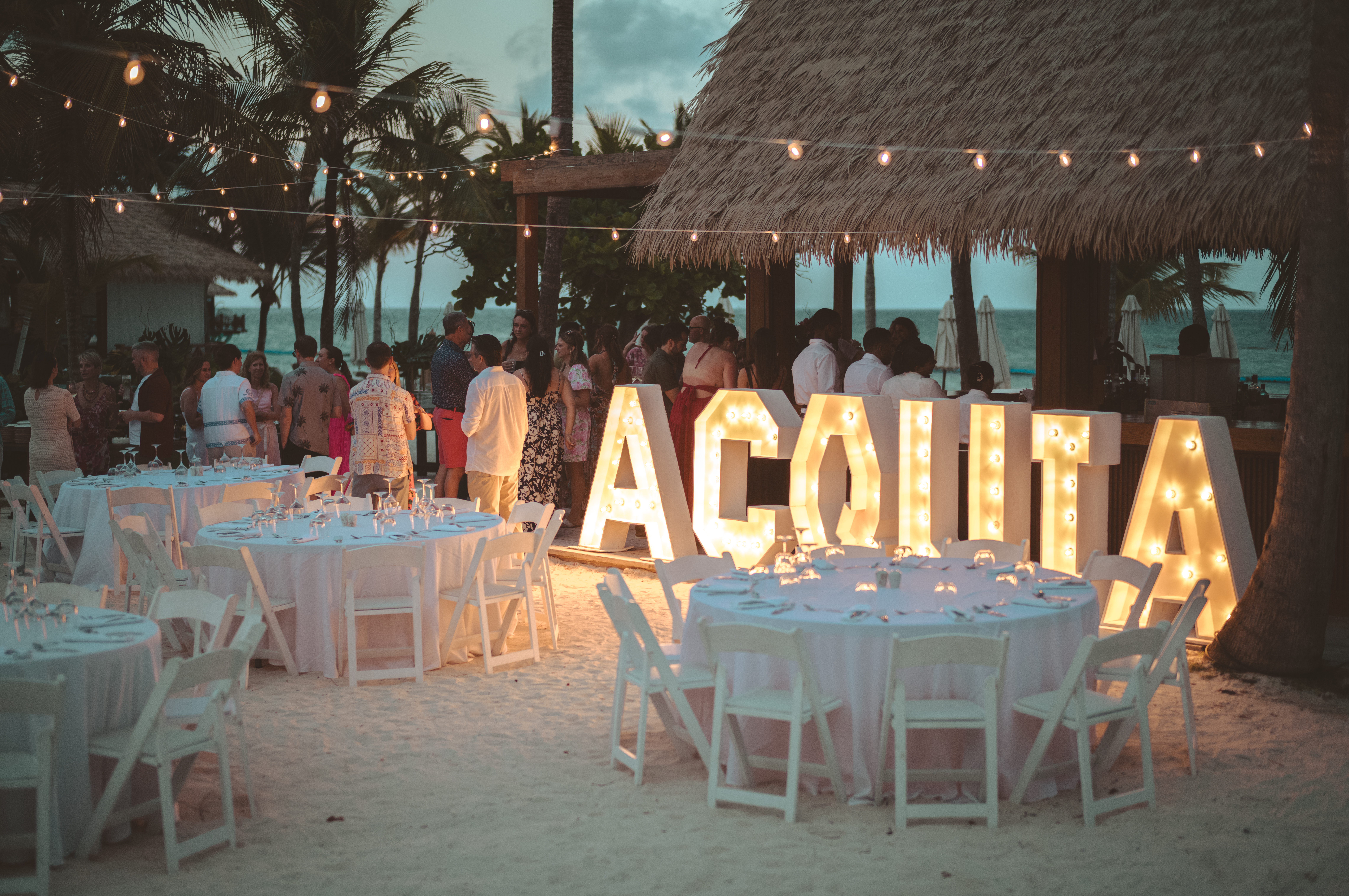 Large illuminated letters spelling &quot;Acquia&quot; are displayed next to tables arranged for a beachside dinner.