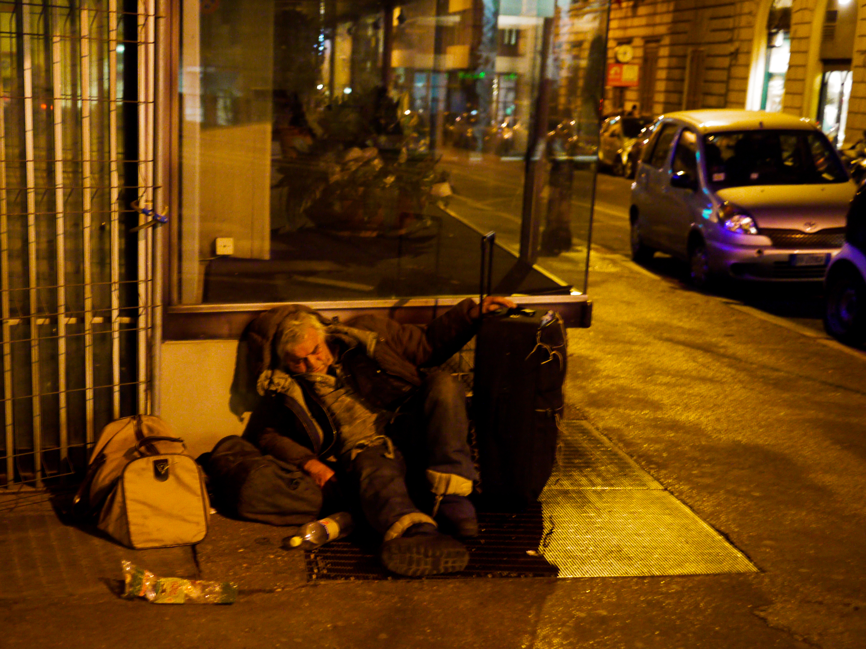 A person is sleeping on the sidewalk, leaning against luggage, with bags and bottles nearby at night.