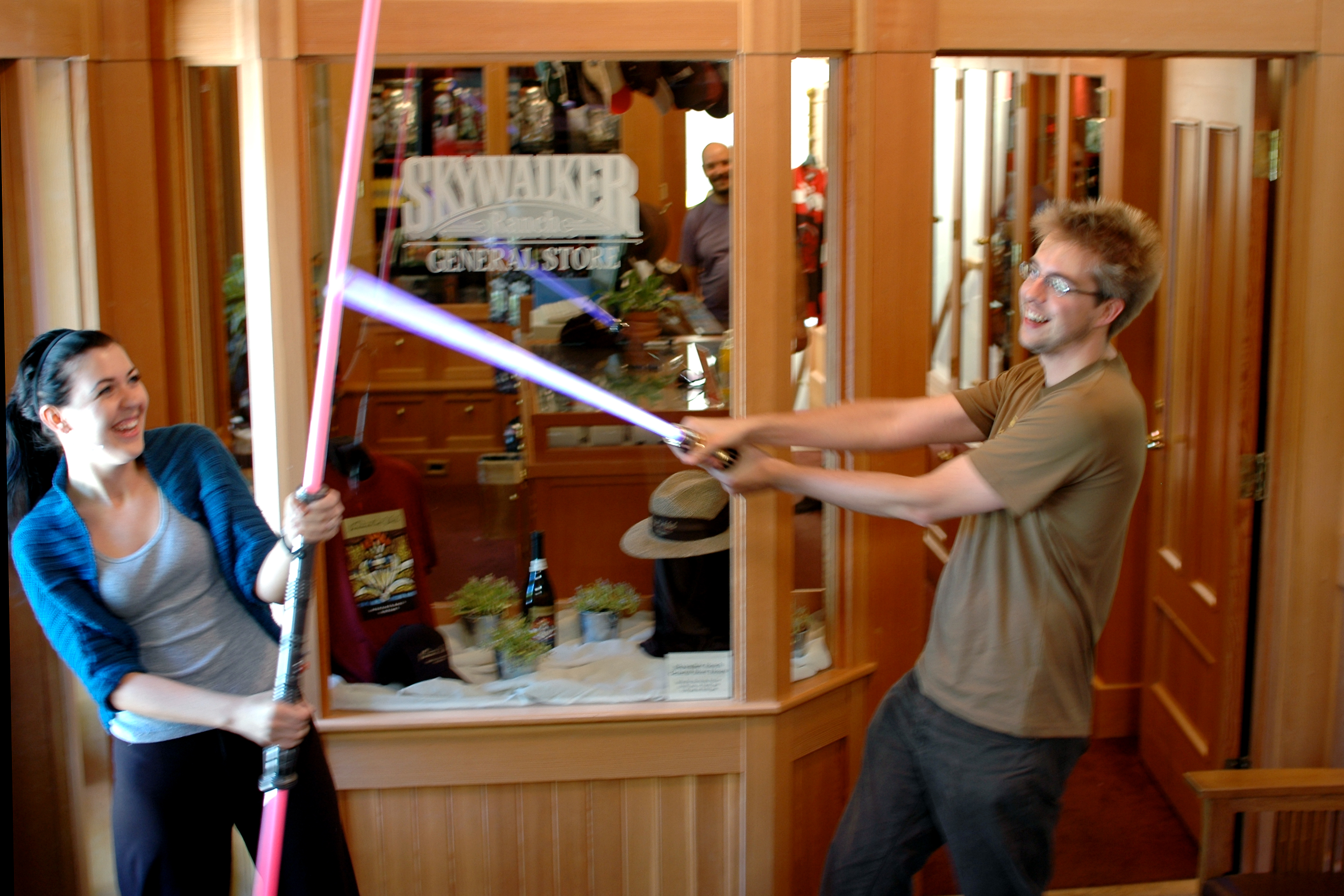 Two people smiling and playfully dueling with toy lightsabers inside a store with a &quot;Skywalker Ranch General Store&quot; sign.