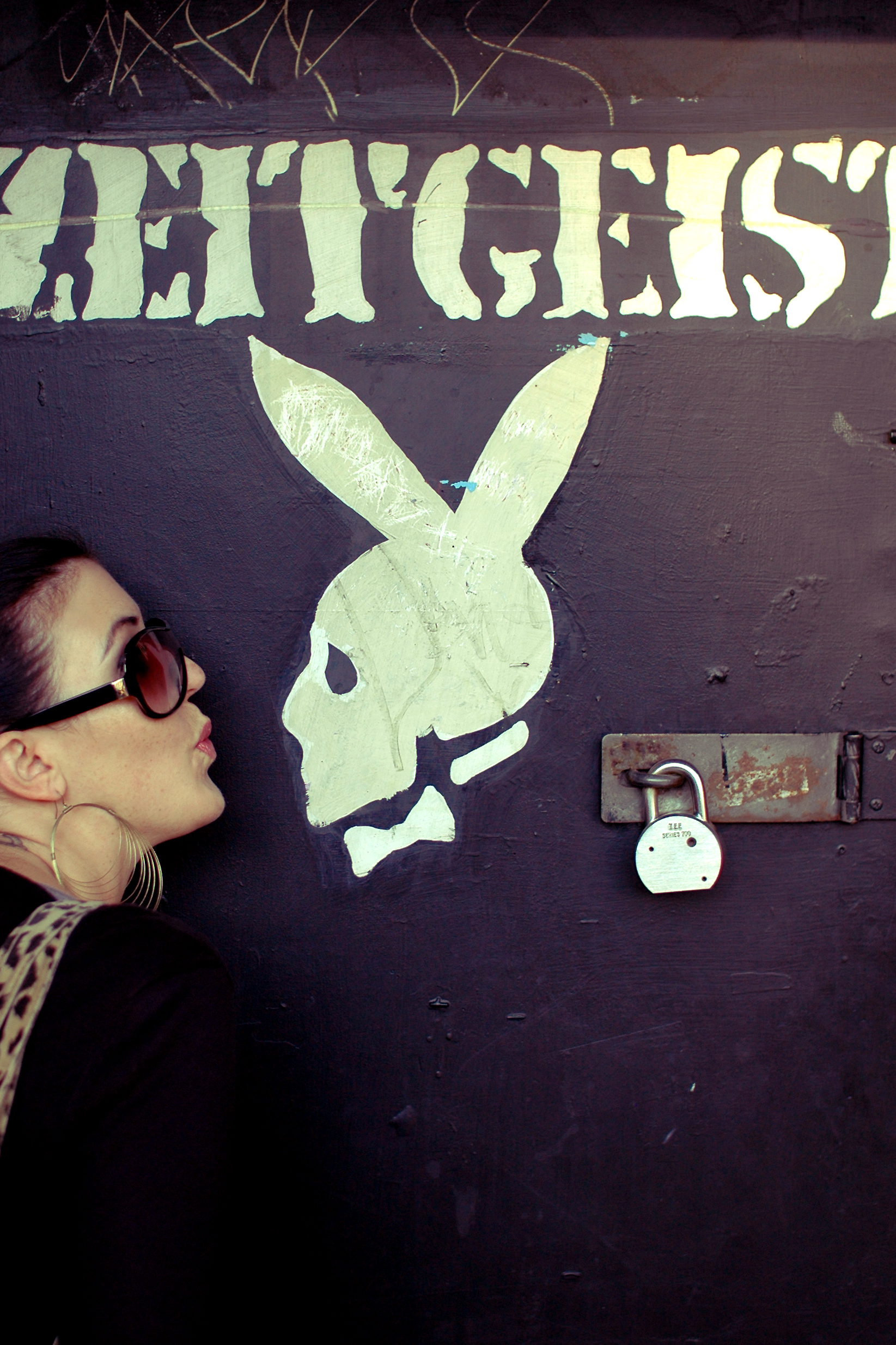 A woman wearing sunglasses playfully kisses a painted skull logo on a dark wall with &quot;Zeitgeist&quot; written above.