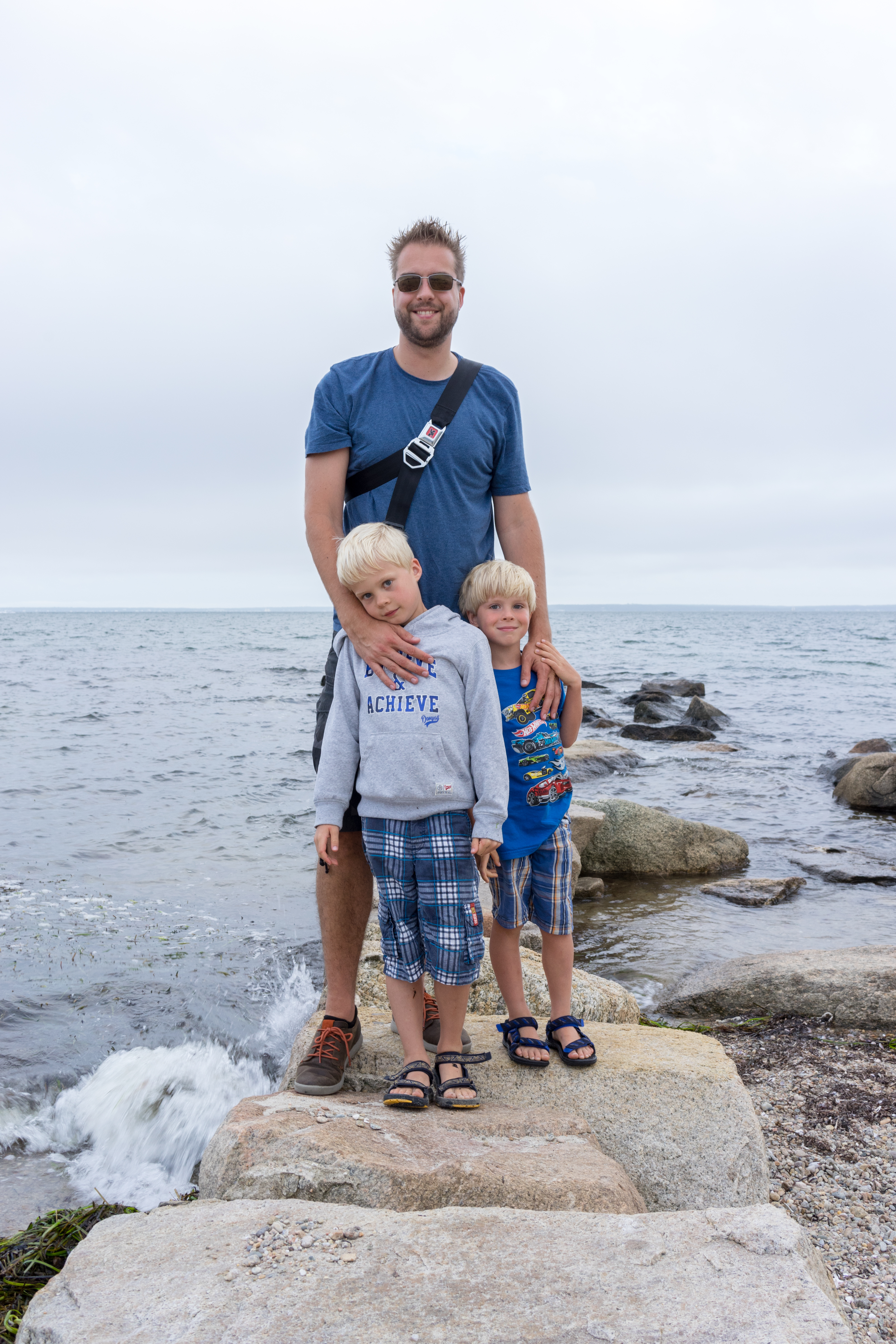 A man stands on rocks by the water with two young children, smiling and posing for a photo.