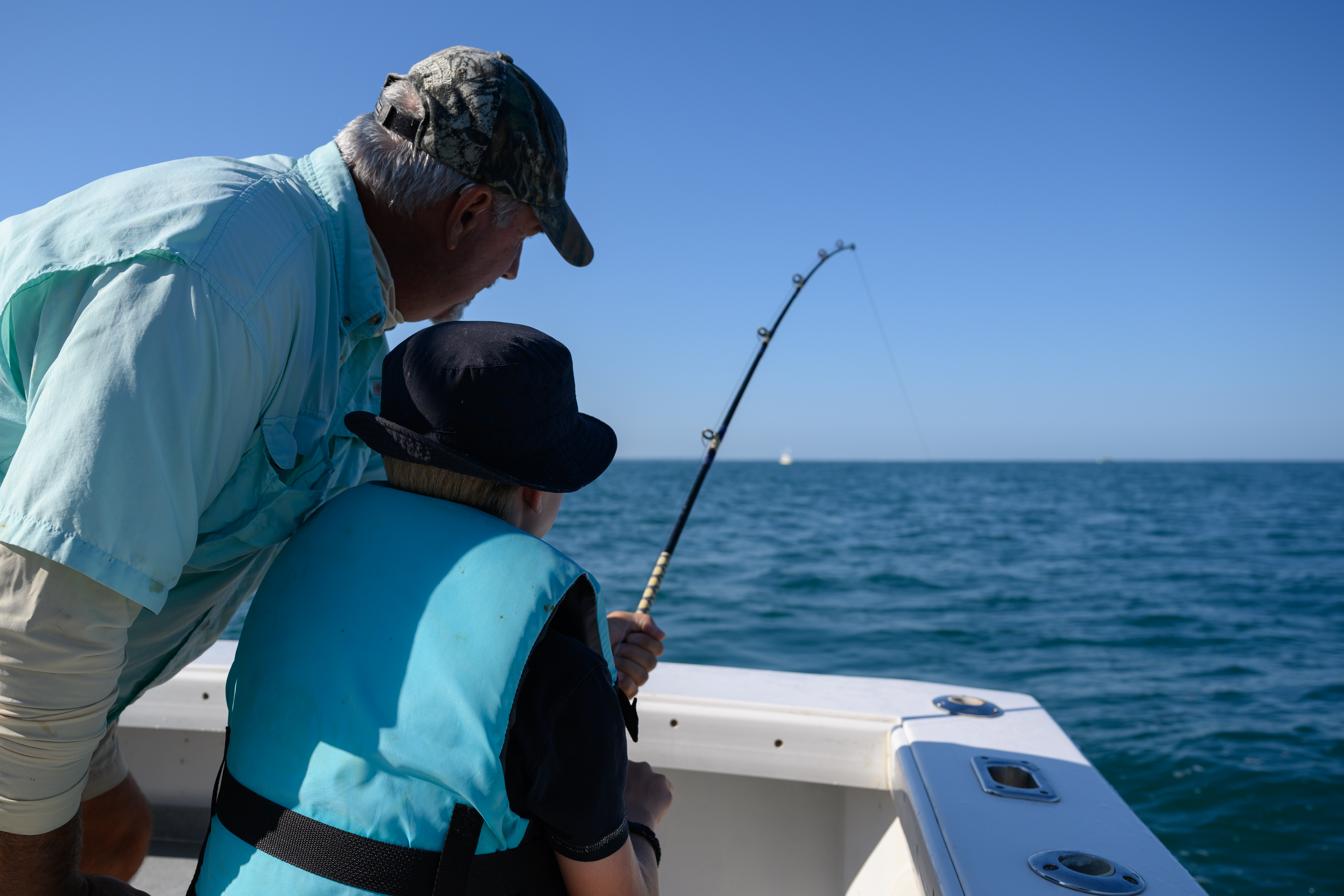 A child wearing a life vest holds a fishing rod while an older man watches on a boat in the ocean.