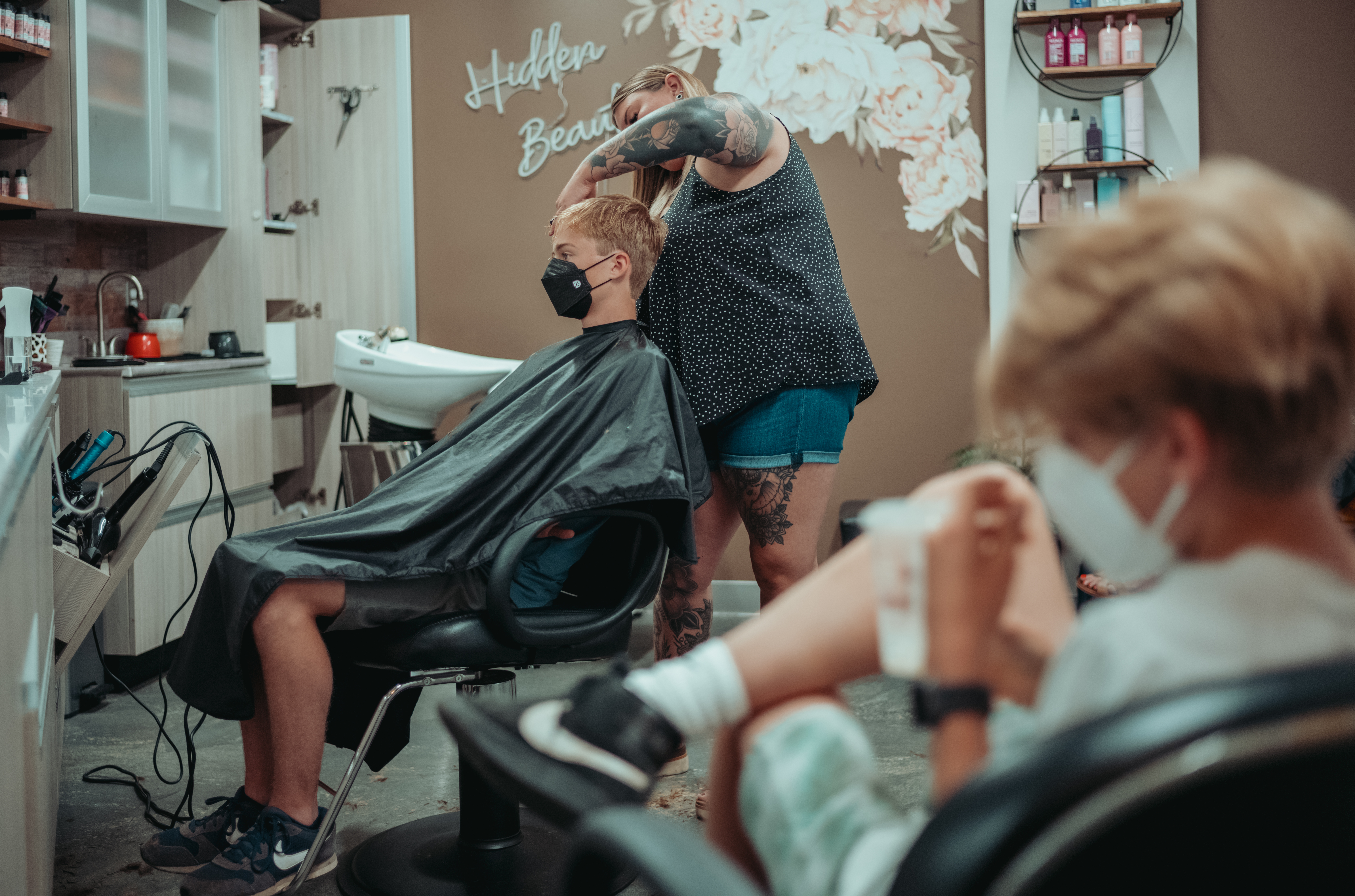 A hairstylist gives a haircut to a seated person wearing a cape, while another person waits nearby.