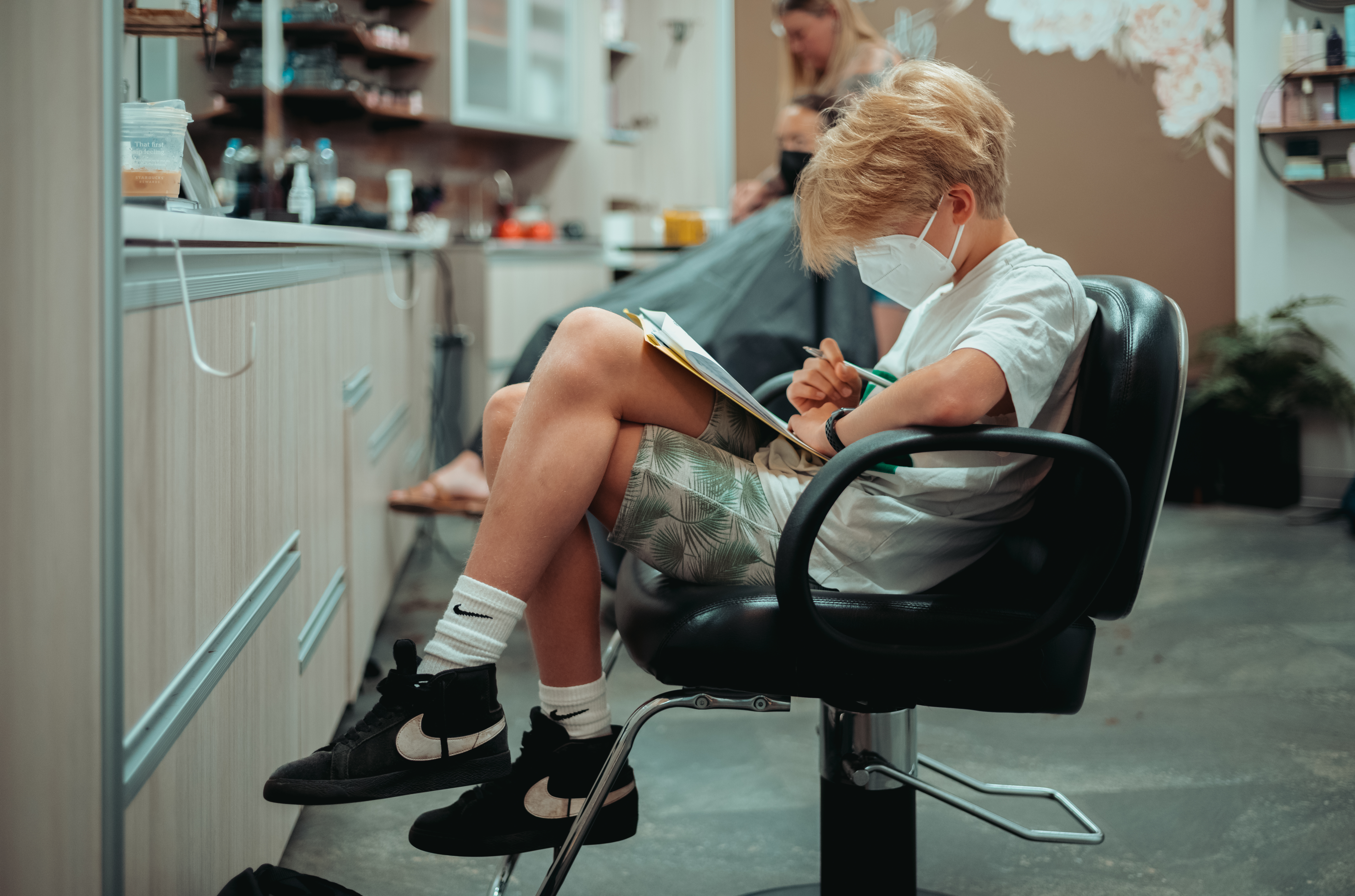 A child wearing a mask sits in a salon chair, drawing in a notebook while waiting for a haircut.