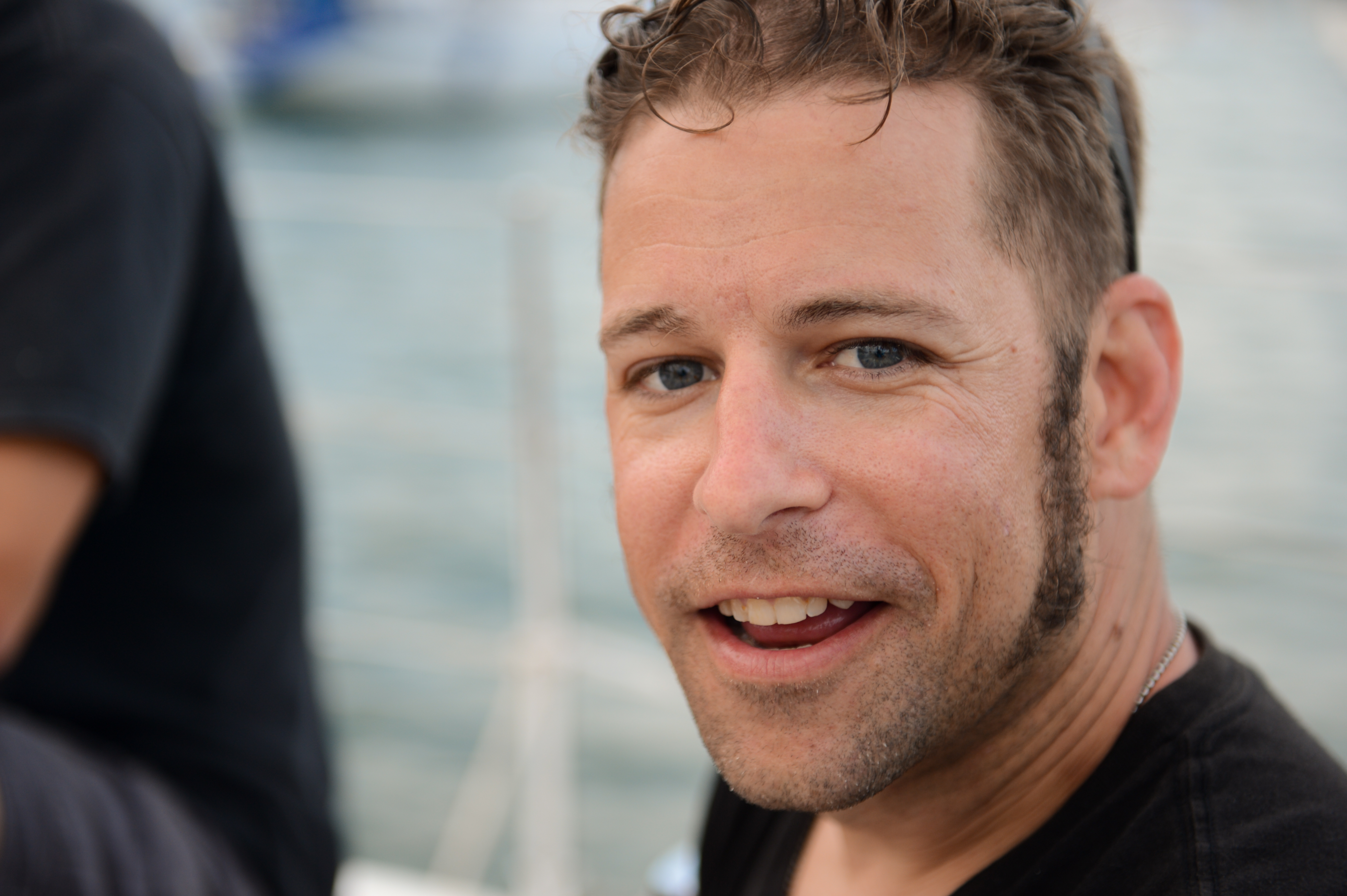 A man with short, curly brown hair, light stubble, and blue eyes smiles at the camera, wearing a black shirt and sunglasses on his head.