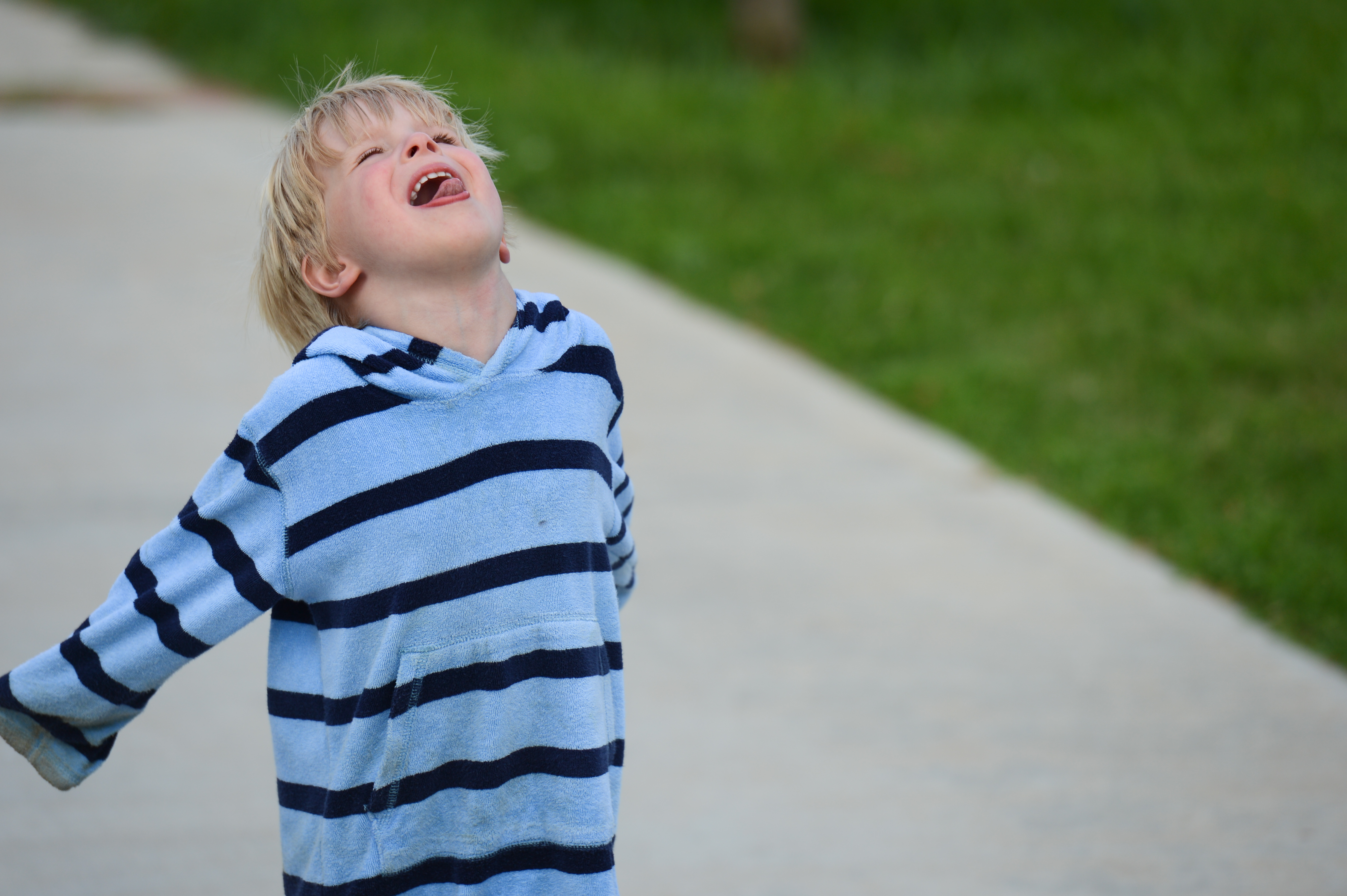 A child in a striped hoodie tilts their head back and smiles while standing on a paved path.