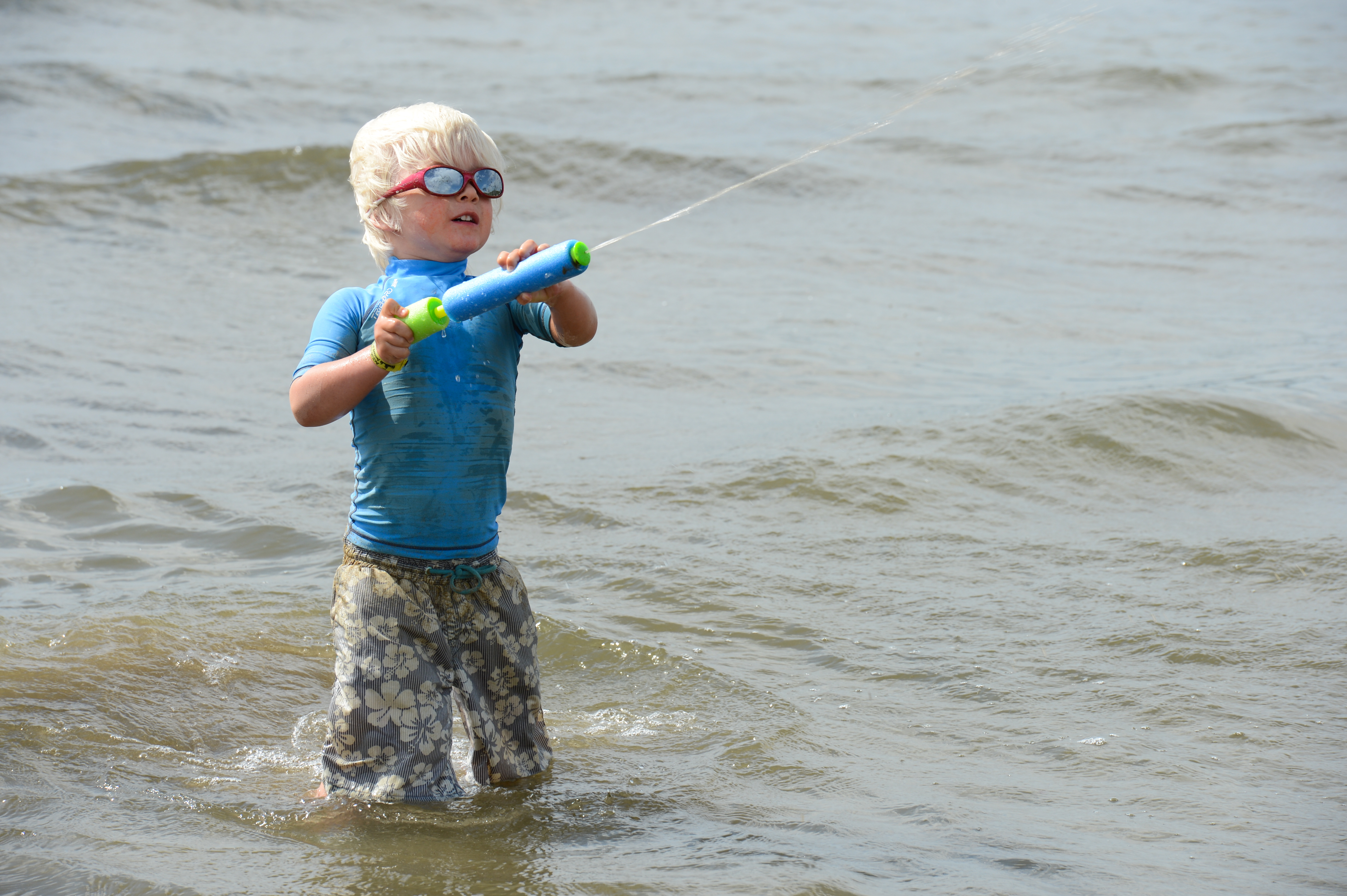 A child wearing sunglasses and a blue shirt plays in the water, spraying a water gun.