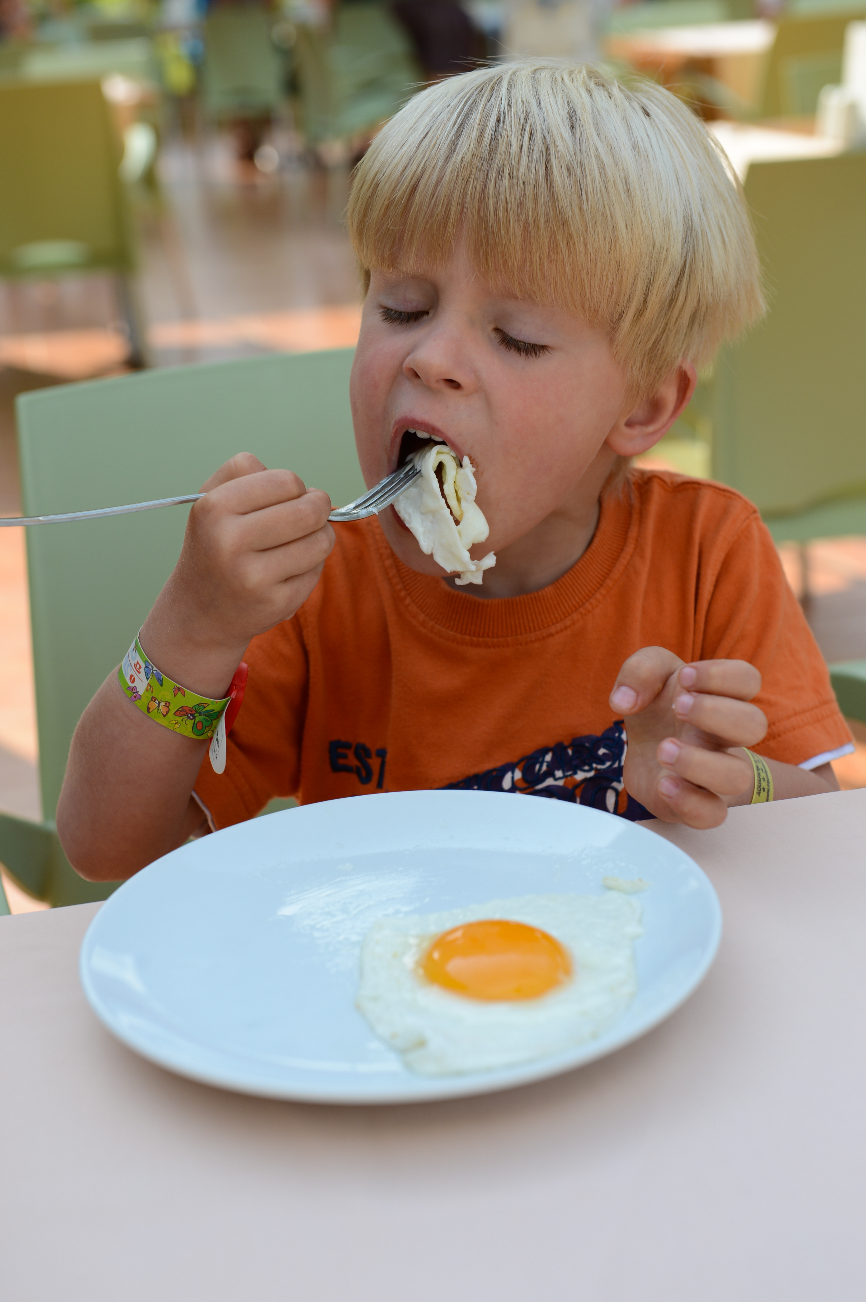 A young child in an orange shirt eats a fried egg with a fork at an outdoor dining area.