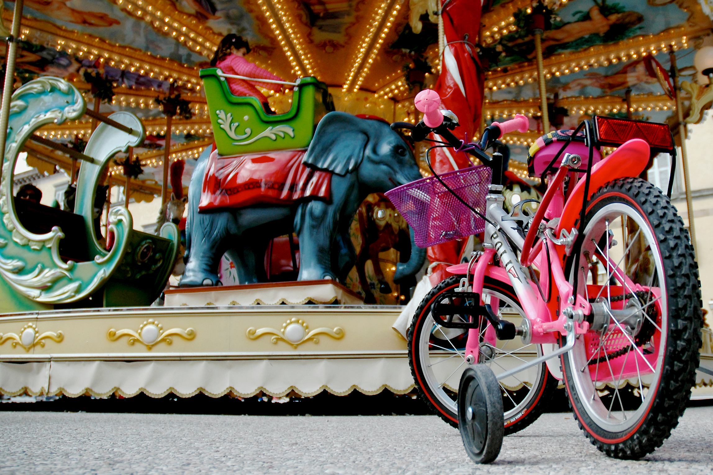 A pink children&#039;s bike with training wheels is parked in front of a carousel where a girl is riding.