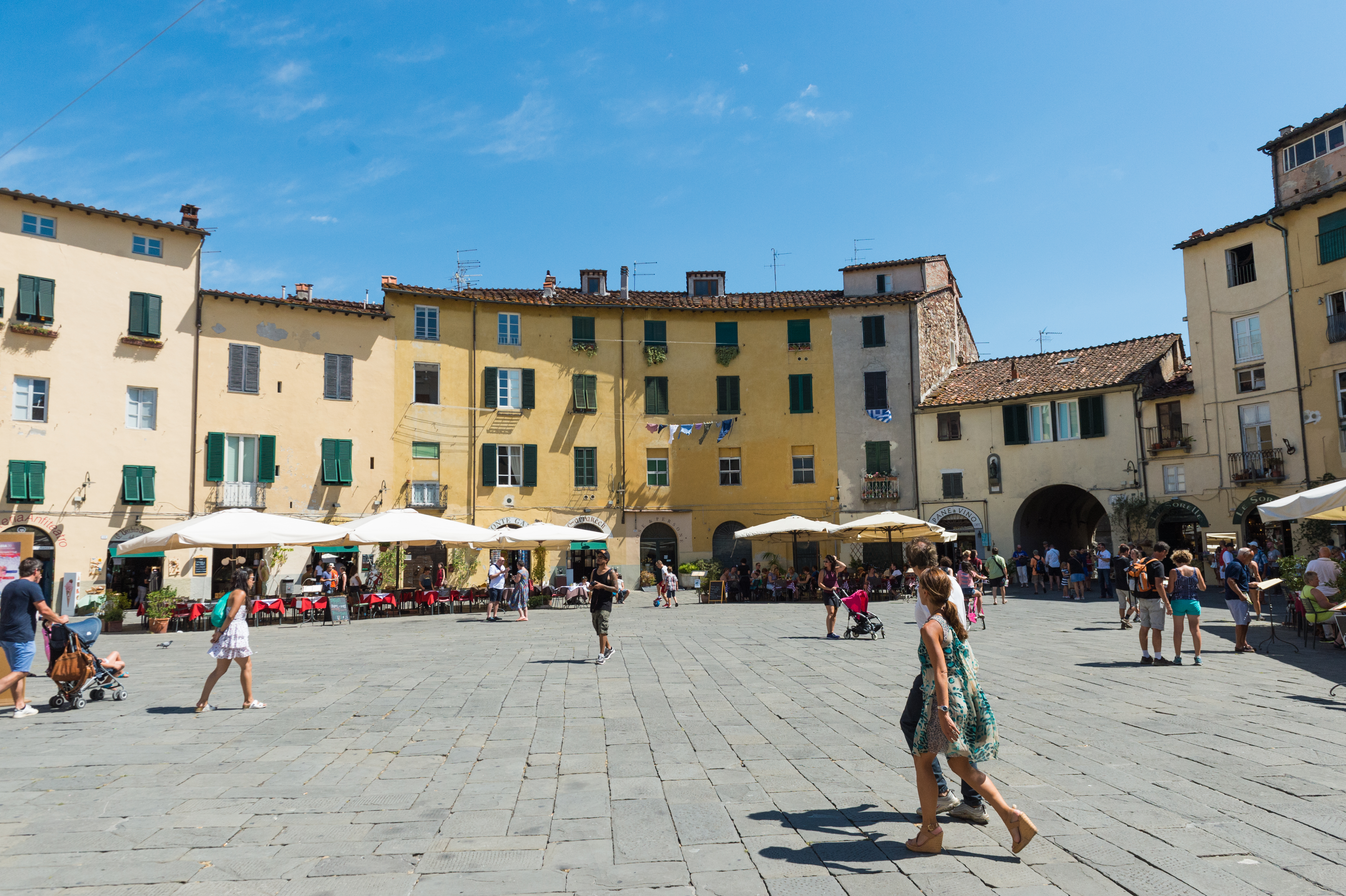 A sunny day at Piazza dell'Anfiteatro in Lucca, with people walking and dining at outdoor cafes.