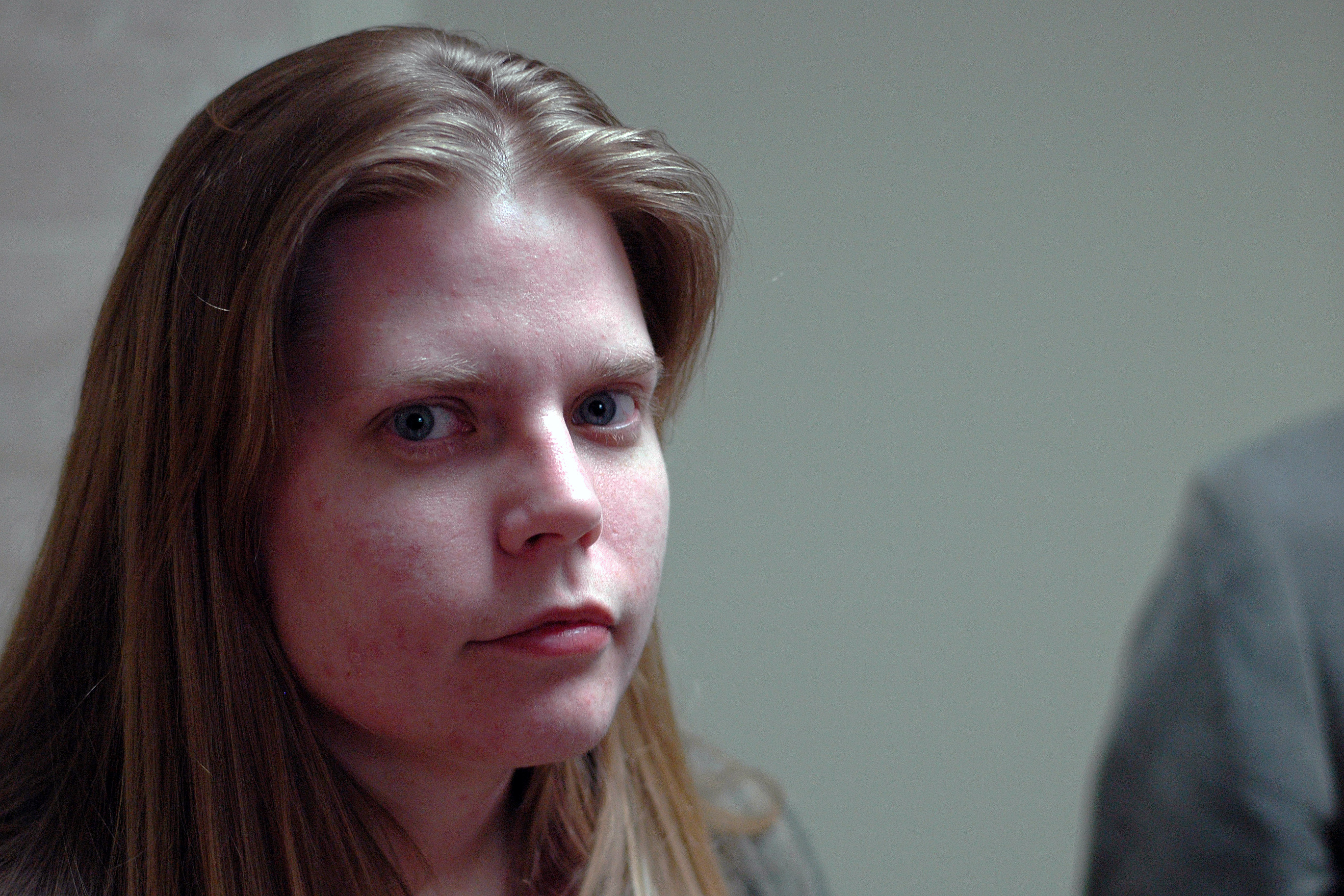 A person with long hair looks directly at the camera with a neutral expression in an indoor setting.
