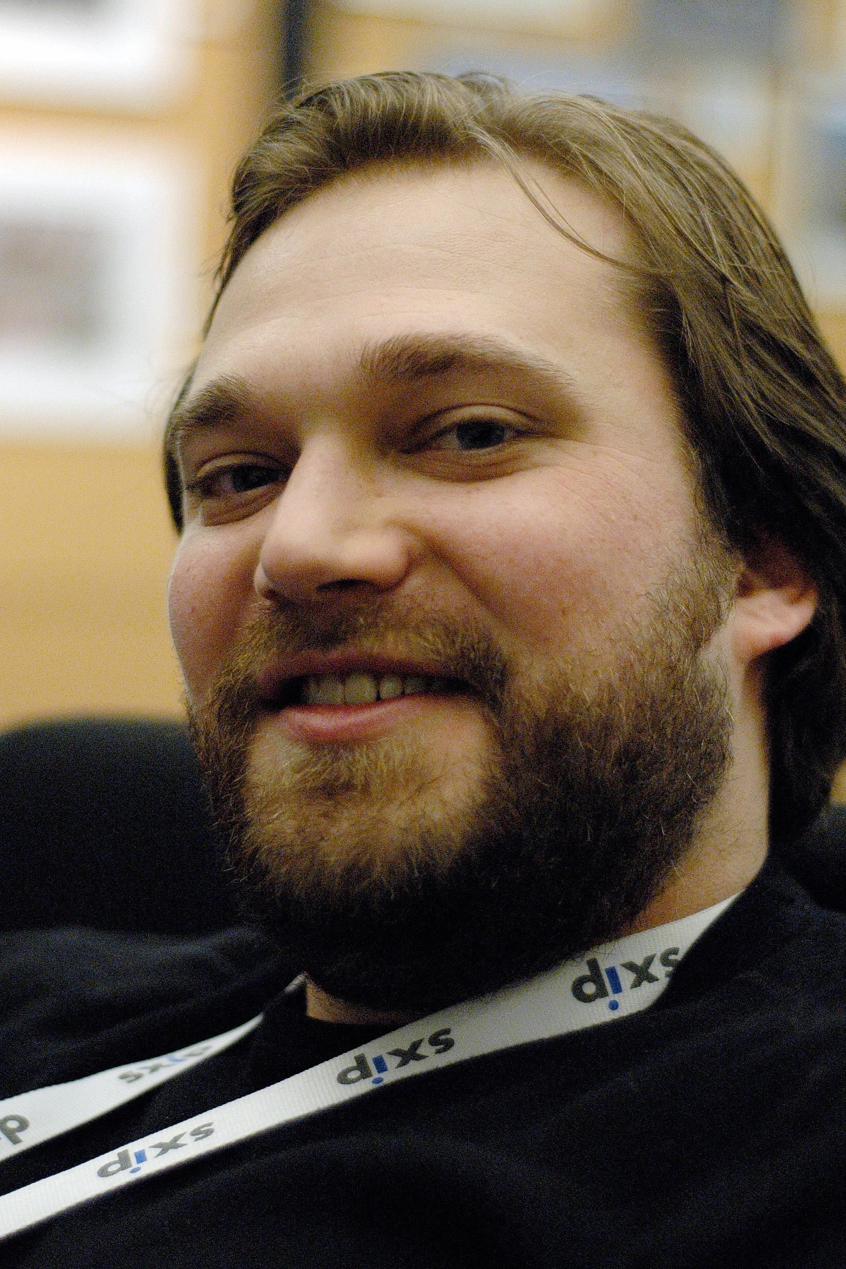 A bearded man wearing a lanyard smiles slightly while looking at the camera in an indoor setting.