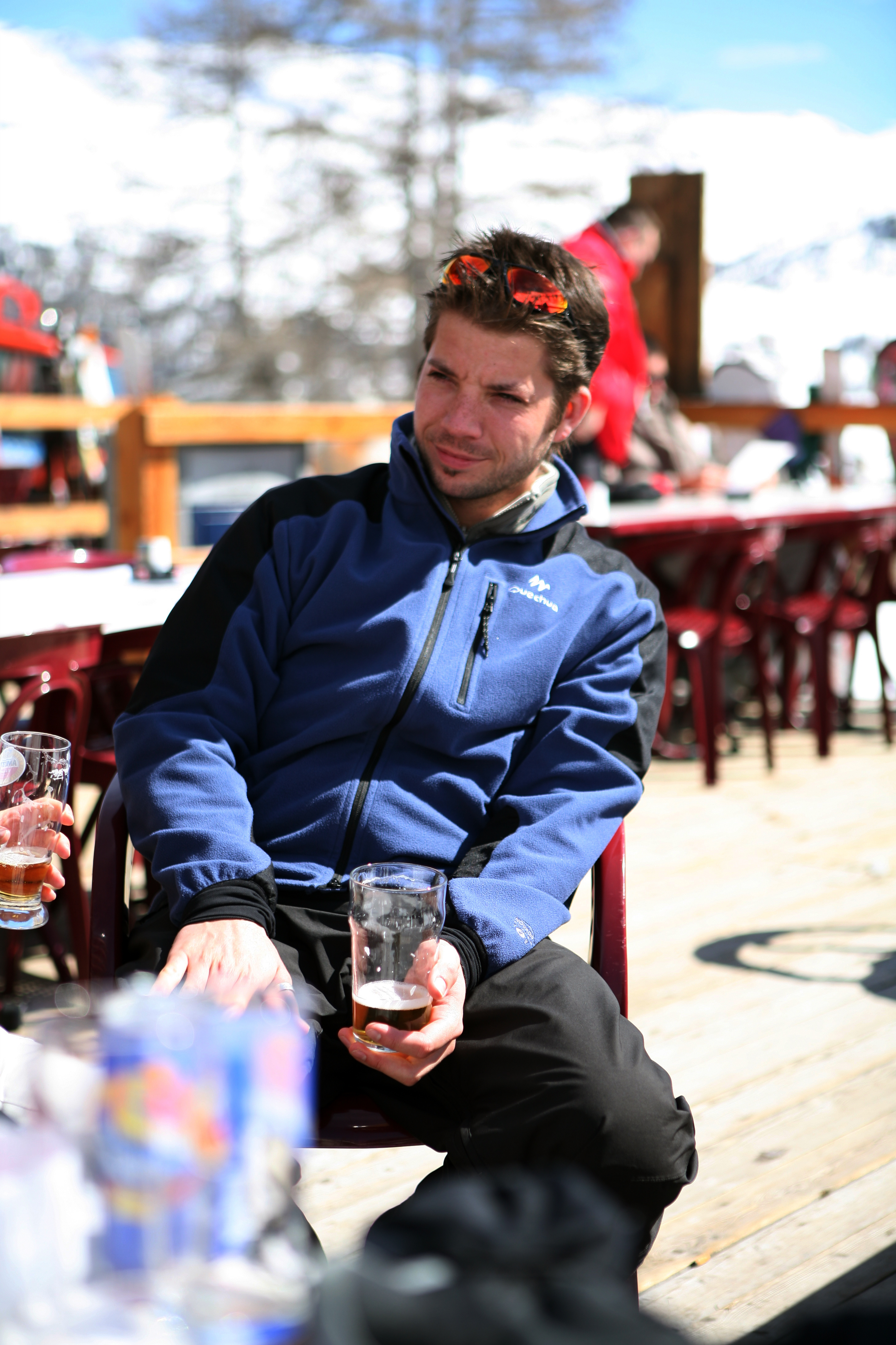 A man in a blue jacket sits outdoors, holding a glass with beer, squinting in the sunlight.
