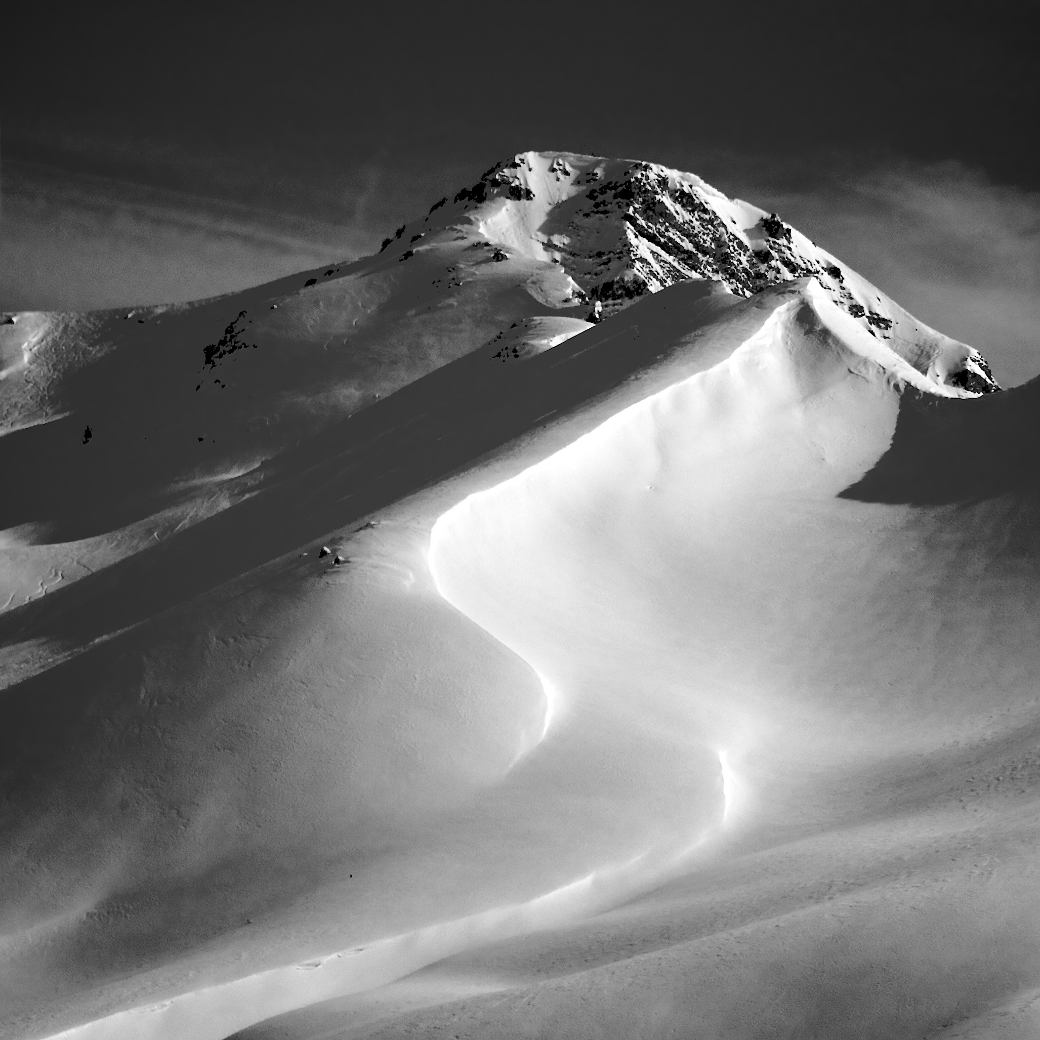 Snow-covered mountain peak with a smooth, curving ski trail leading down the slope under a dark sky.