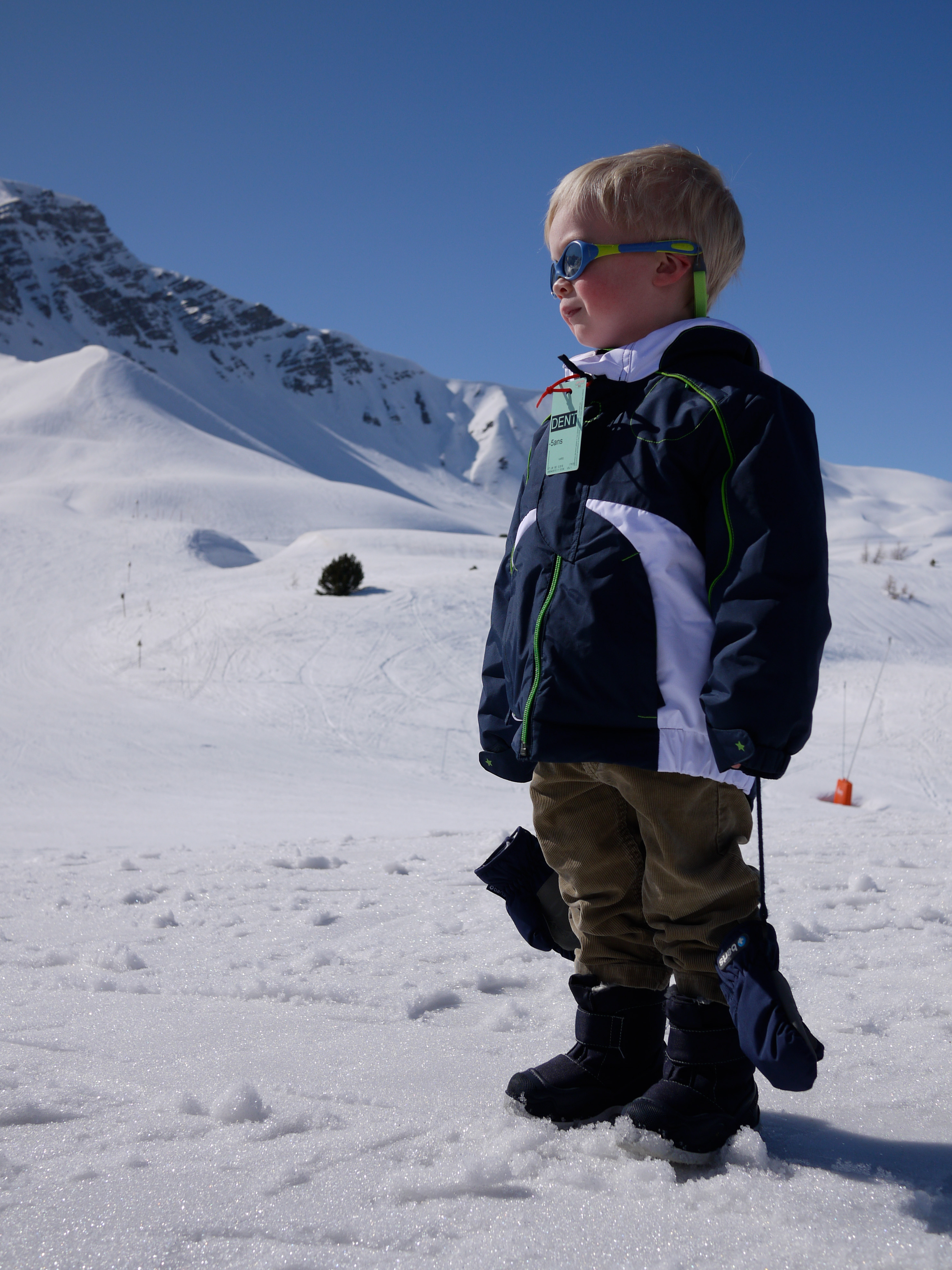 A young child wearing a winter jacket and goggles stands on a snowy slope, looking into the distance.