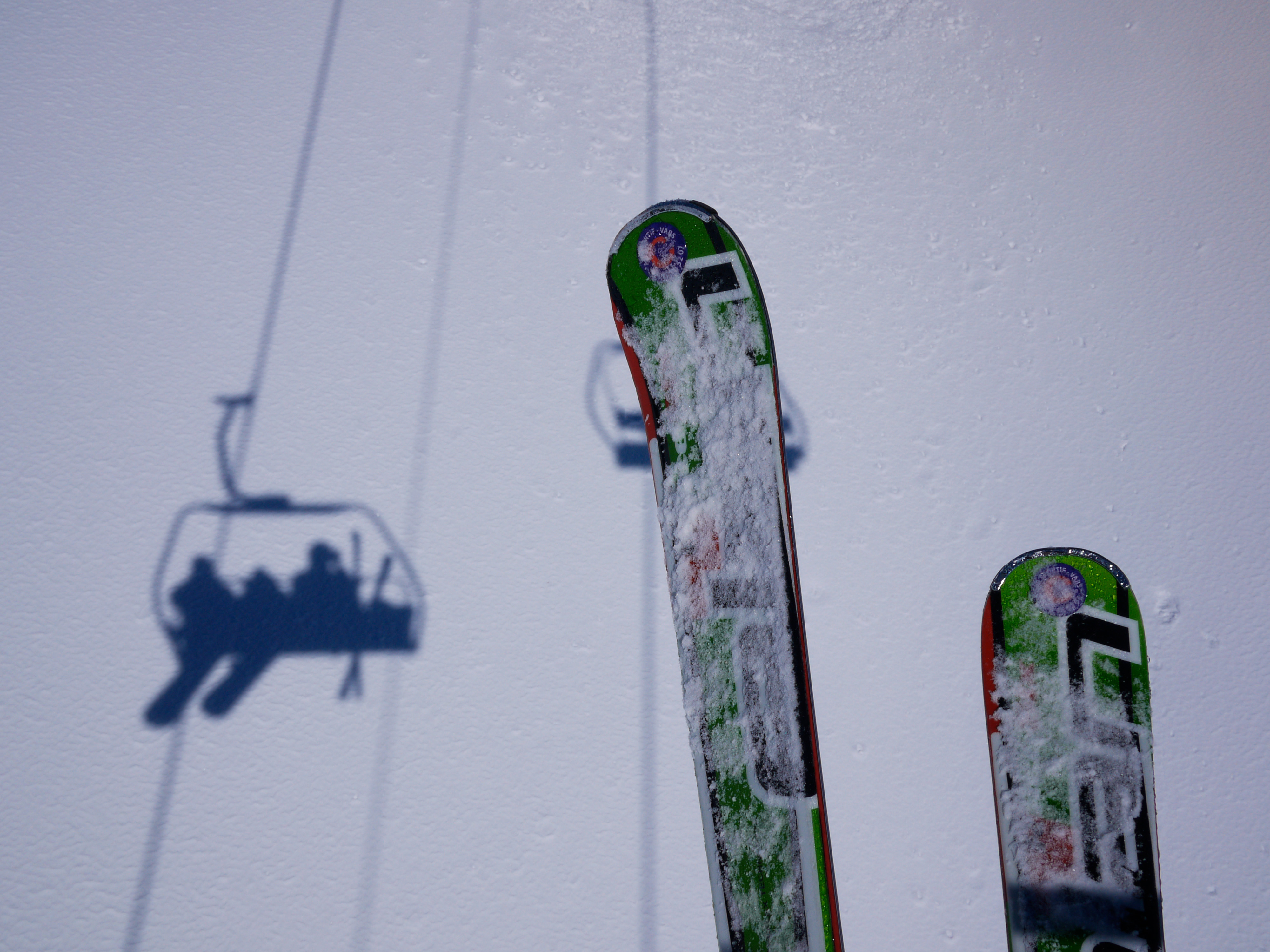 A pair of snow-covered skis with a ski lift shadow on the snowy ground.