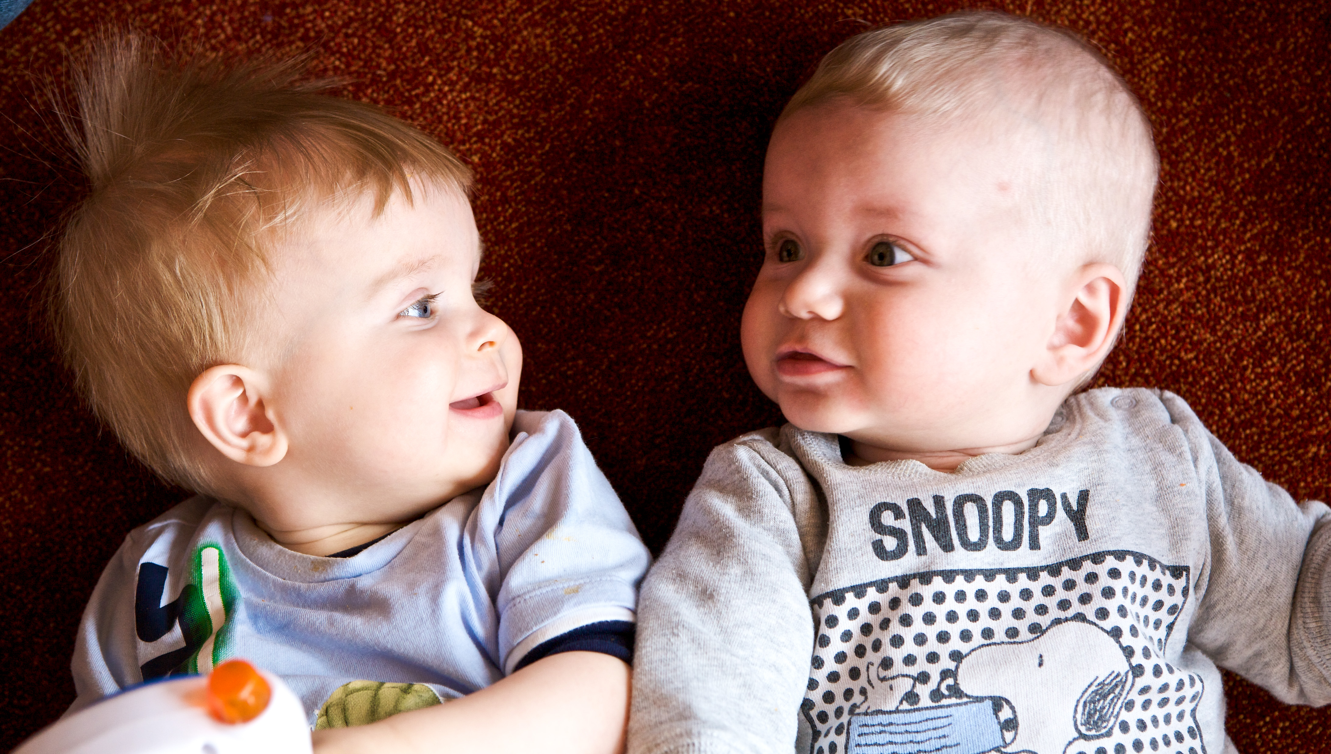 Two babies lie on a red carpet, facing each other.