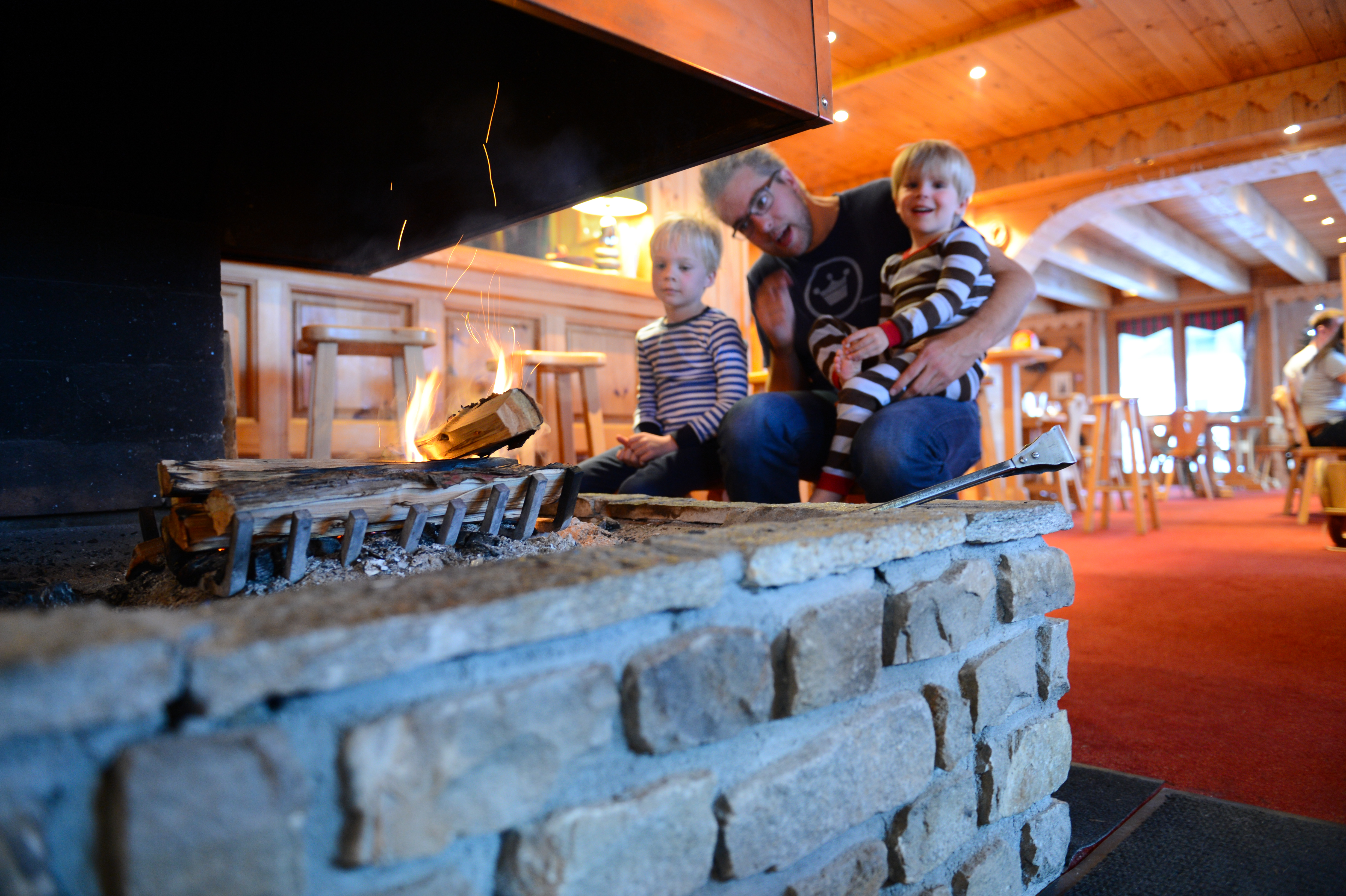 A man and two children sit near a stone fireplace with a burning fire in a cozy wooden room.