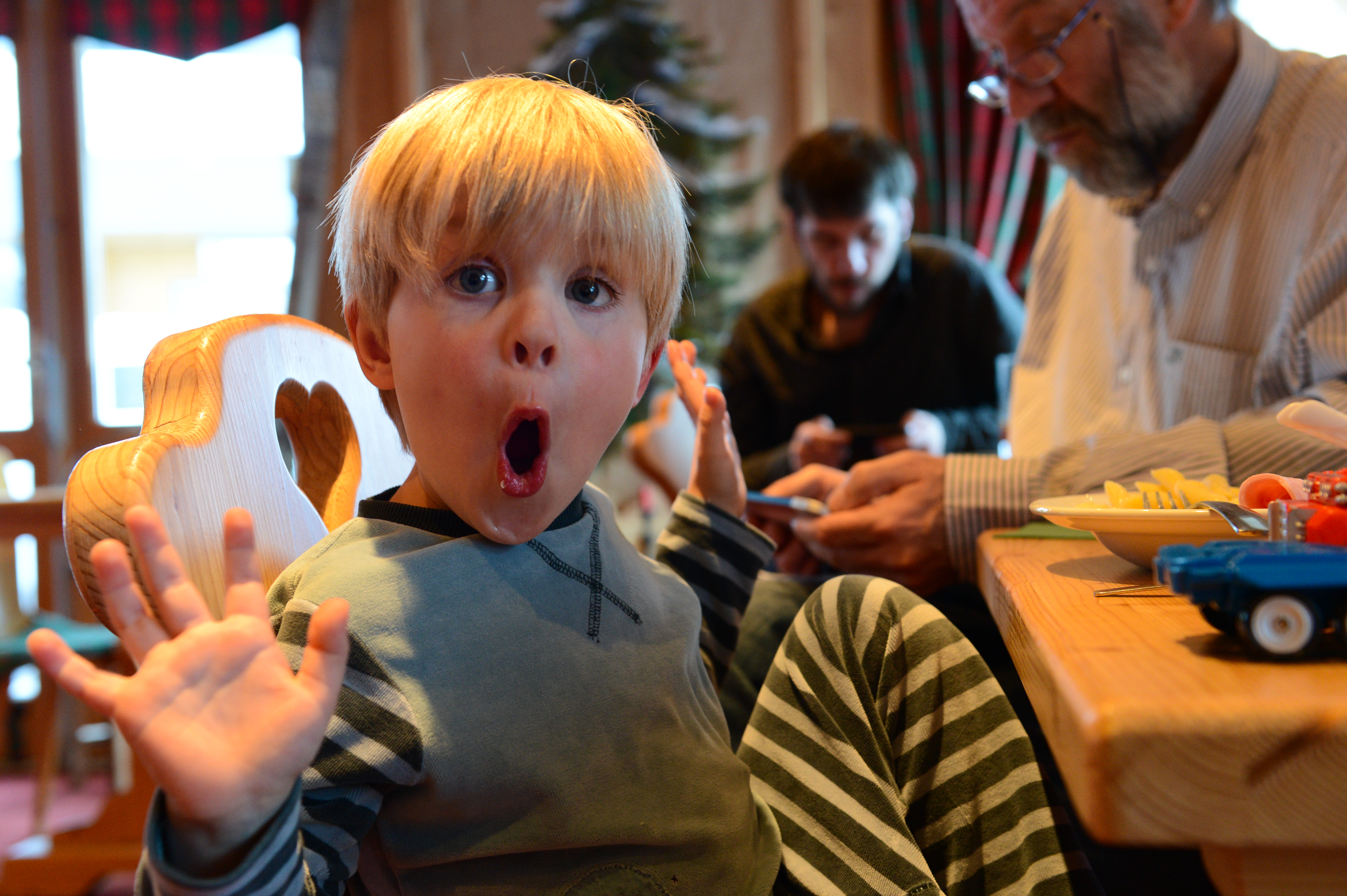 A young child with blond hair makes a surprised face, hands raised, while sitting at a table with adults.