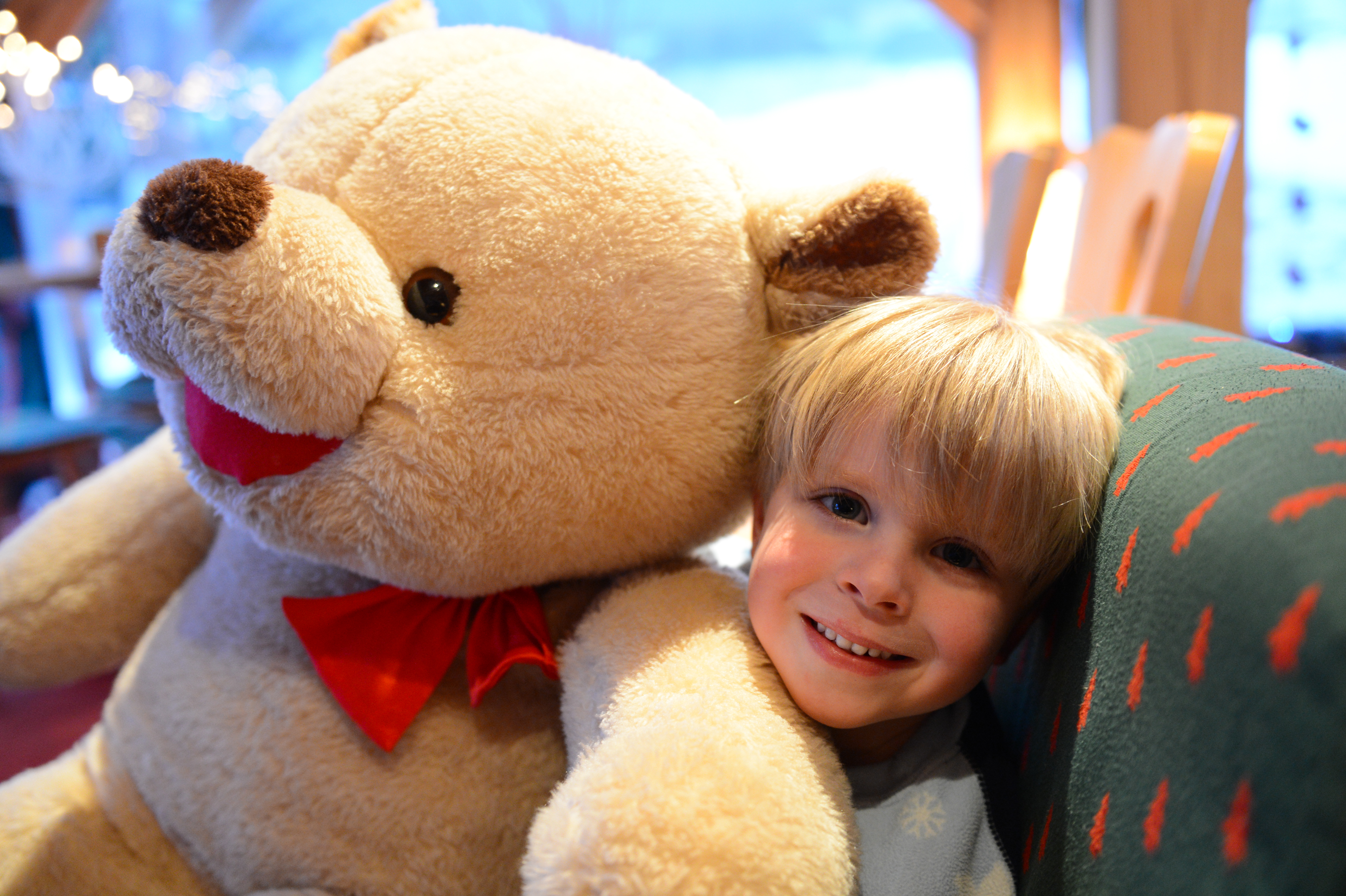 A smiling child sits next to a large teddy bear with a red bow, leaning against a couch.