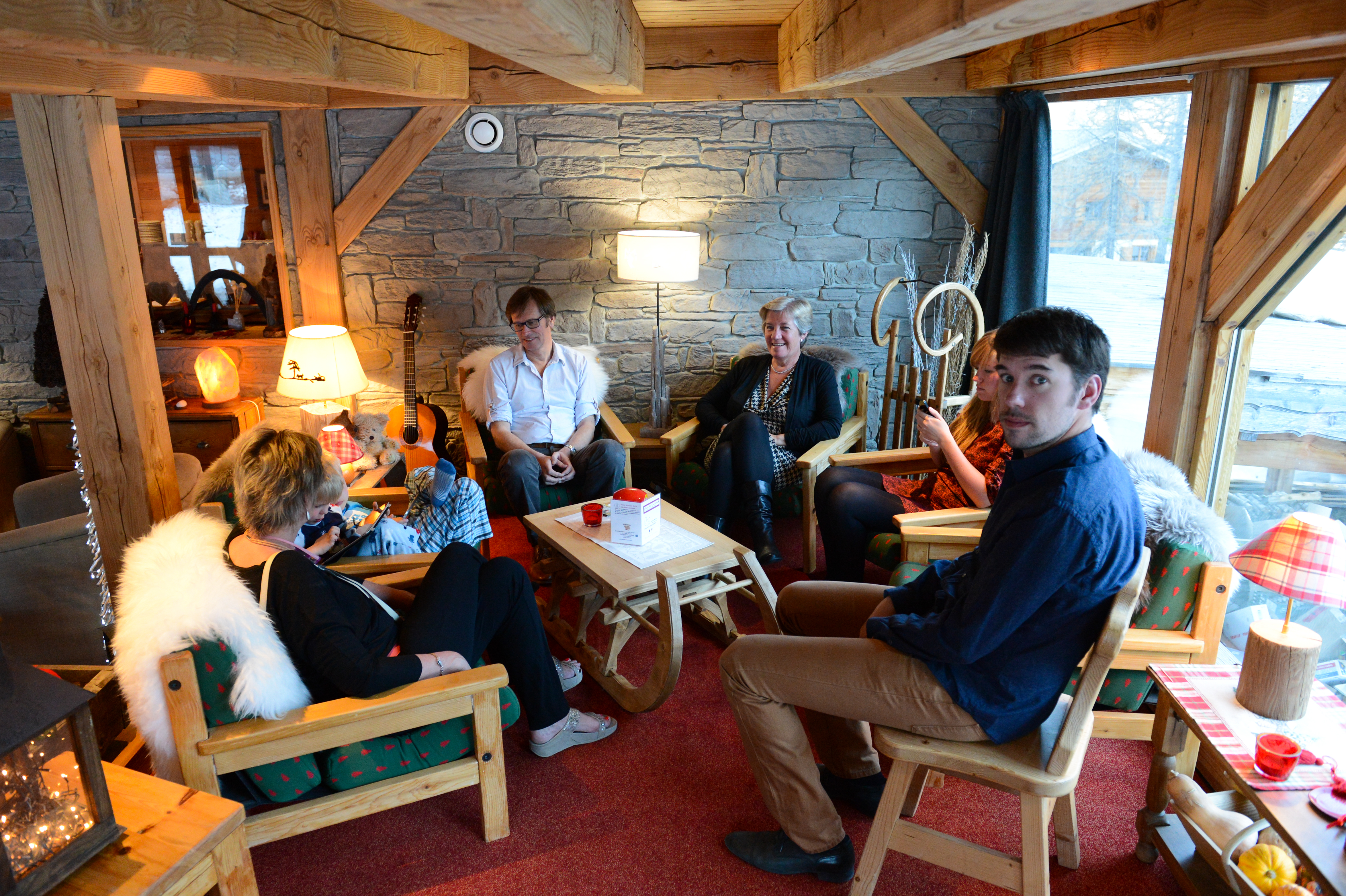 A group of people sits in a cozy wooden cabin, chatting and relaxing around a small table.
