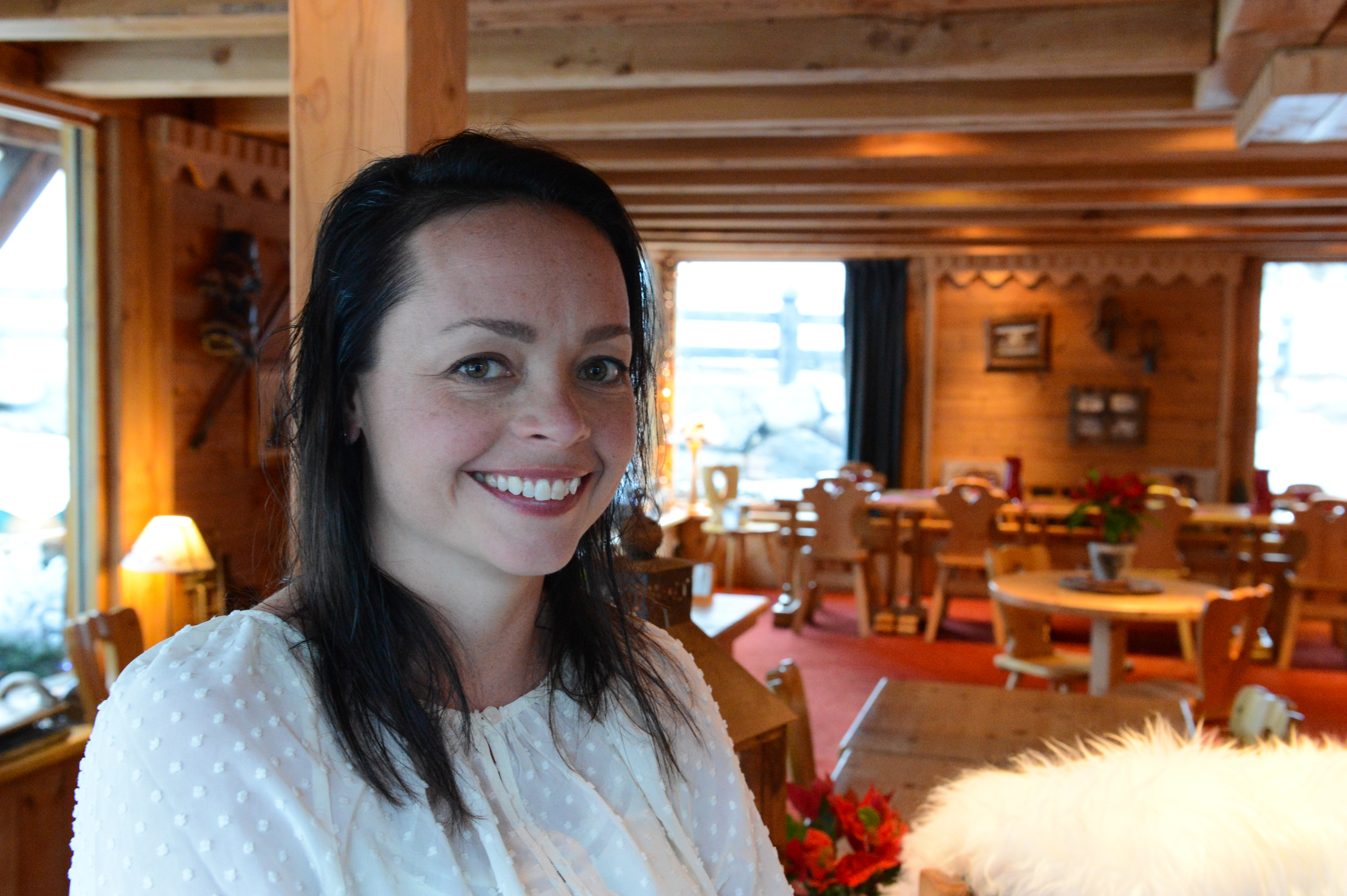 A woman in a white blouse smiles at the camera inside a cozy wooden restaurant or lodge.