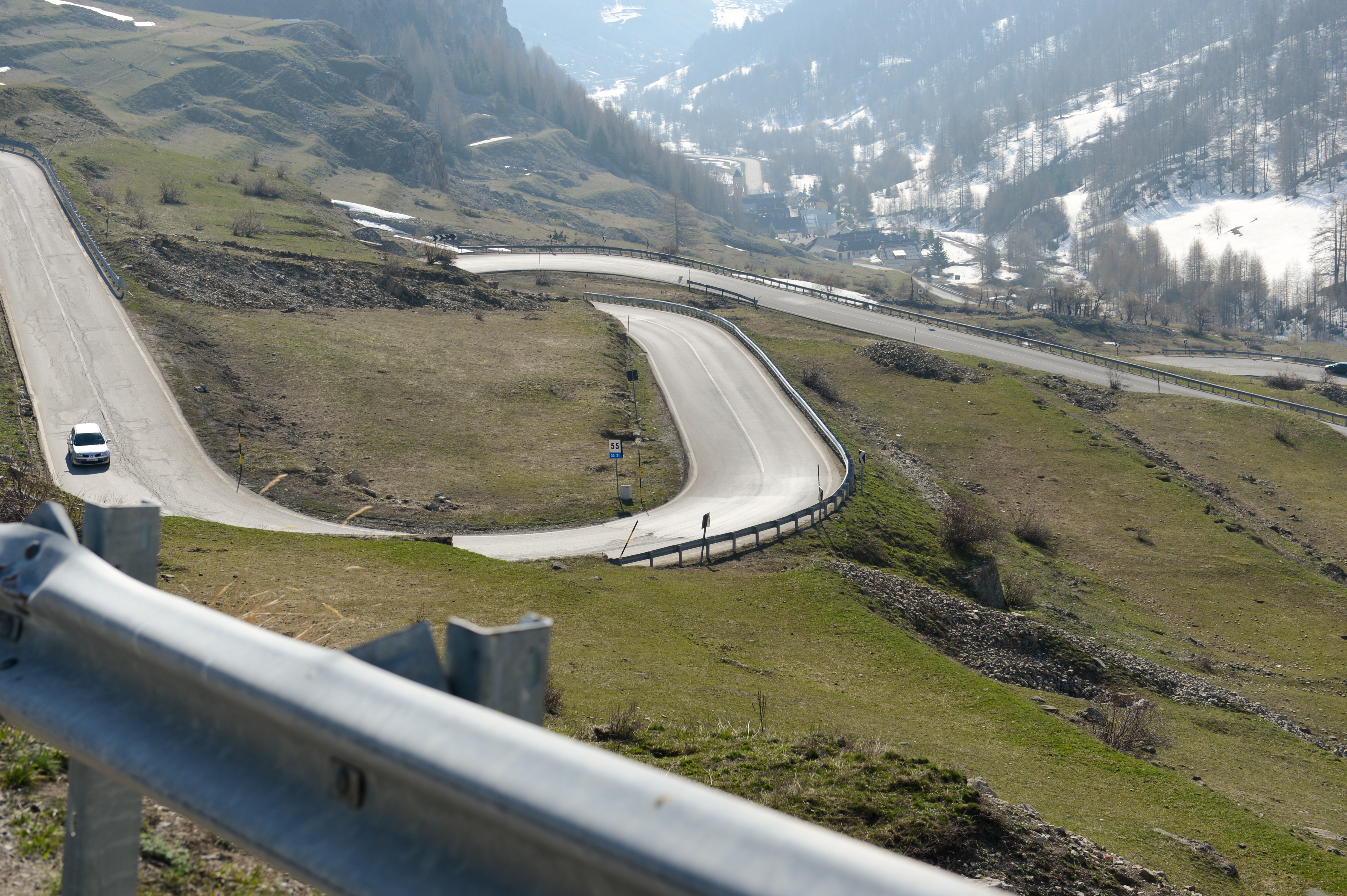 A winding mountain road with a white car driving downhill, surrounded by grassy slopes and patches of snow.