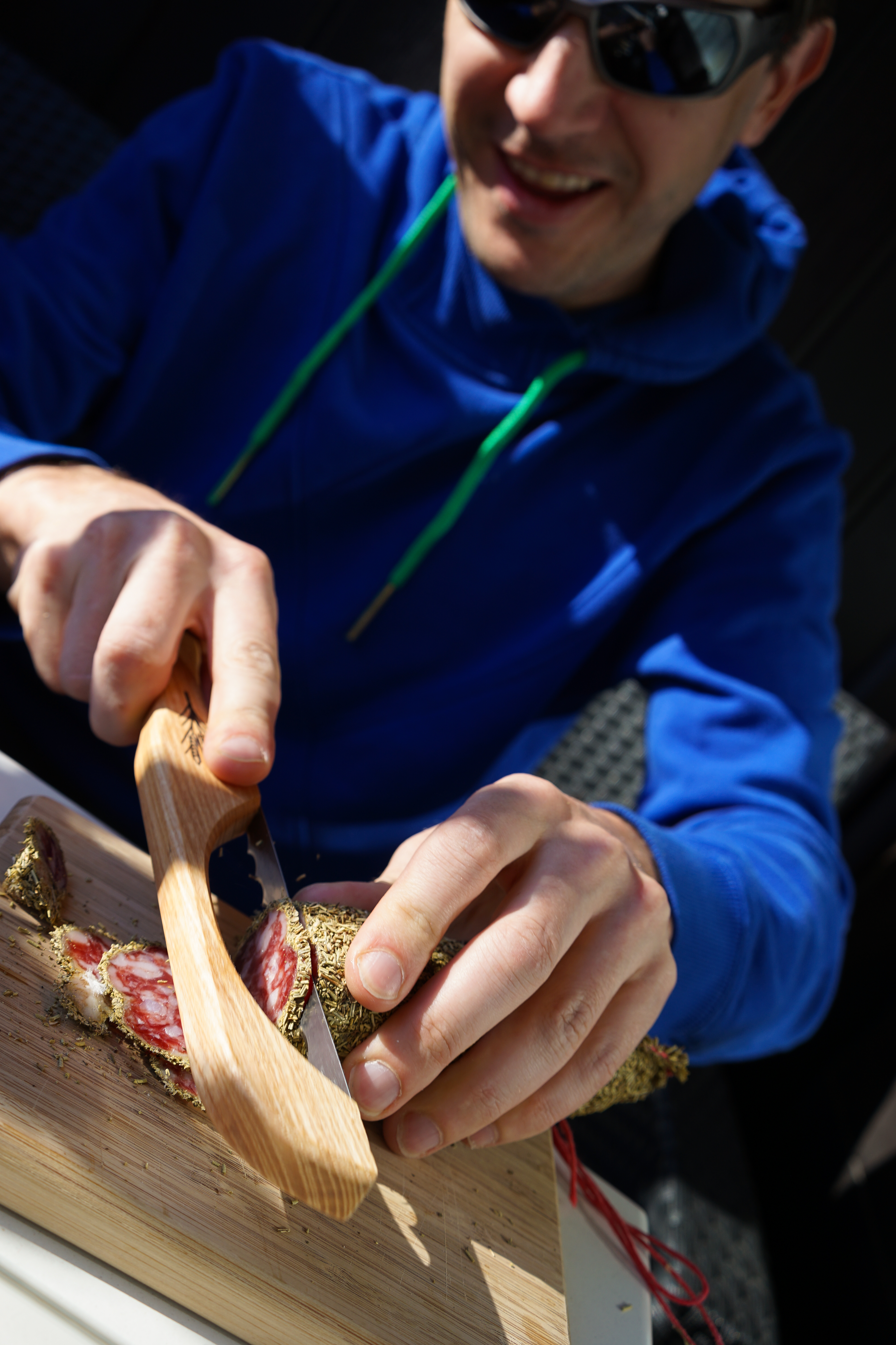 A person in a blue hoodie slices a herb-crusted sausage on a wooden cutting board using a special knife.
