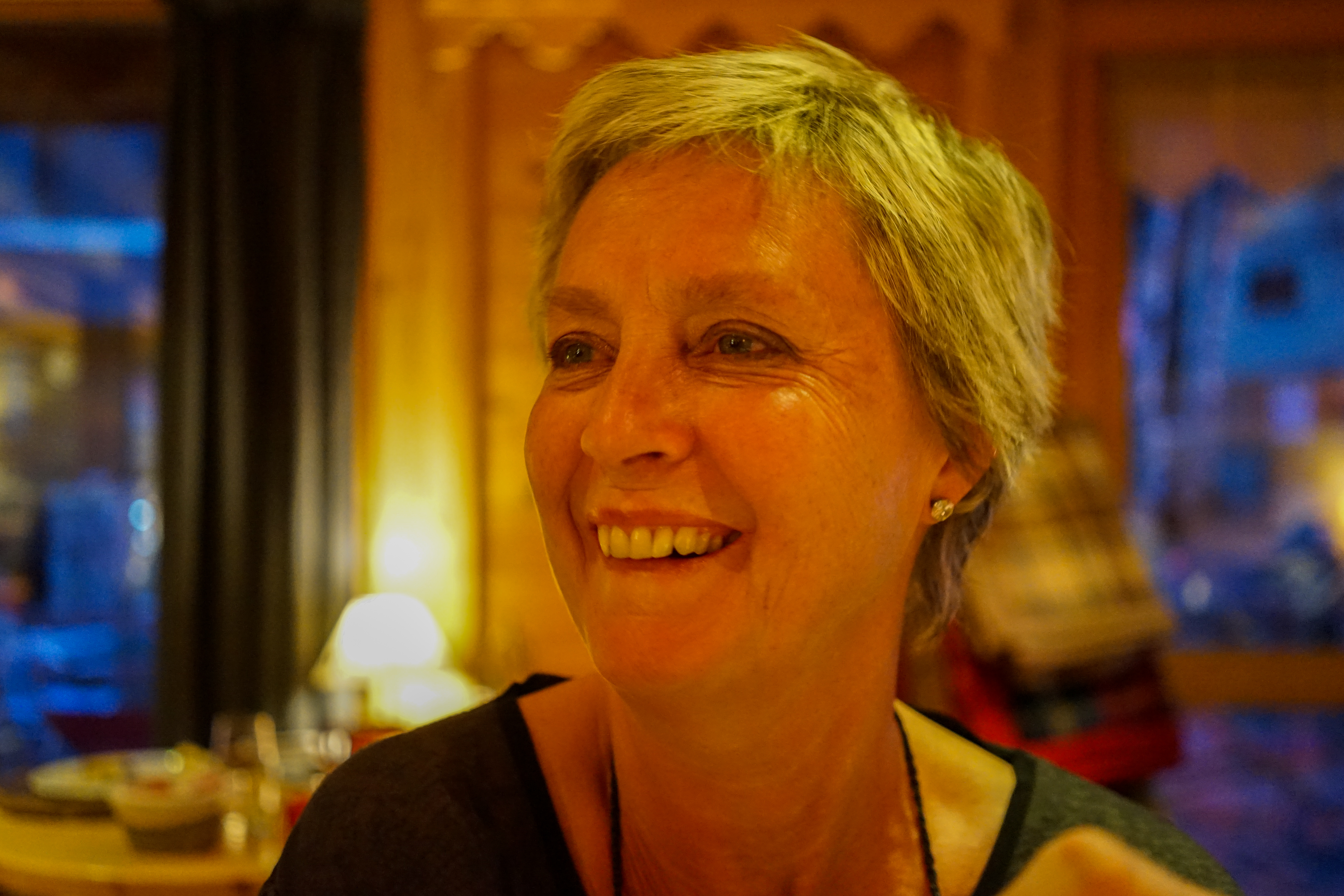 A woman with short hair smiles warmly in a dimly lit indoor setting.