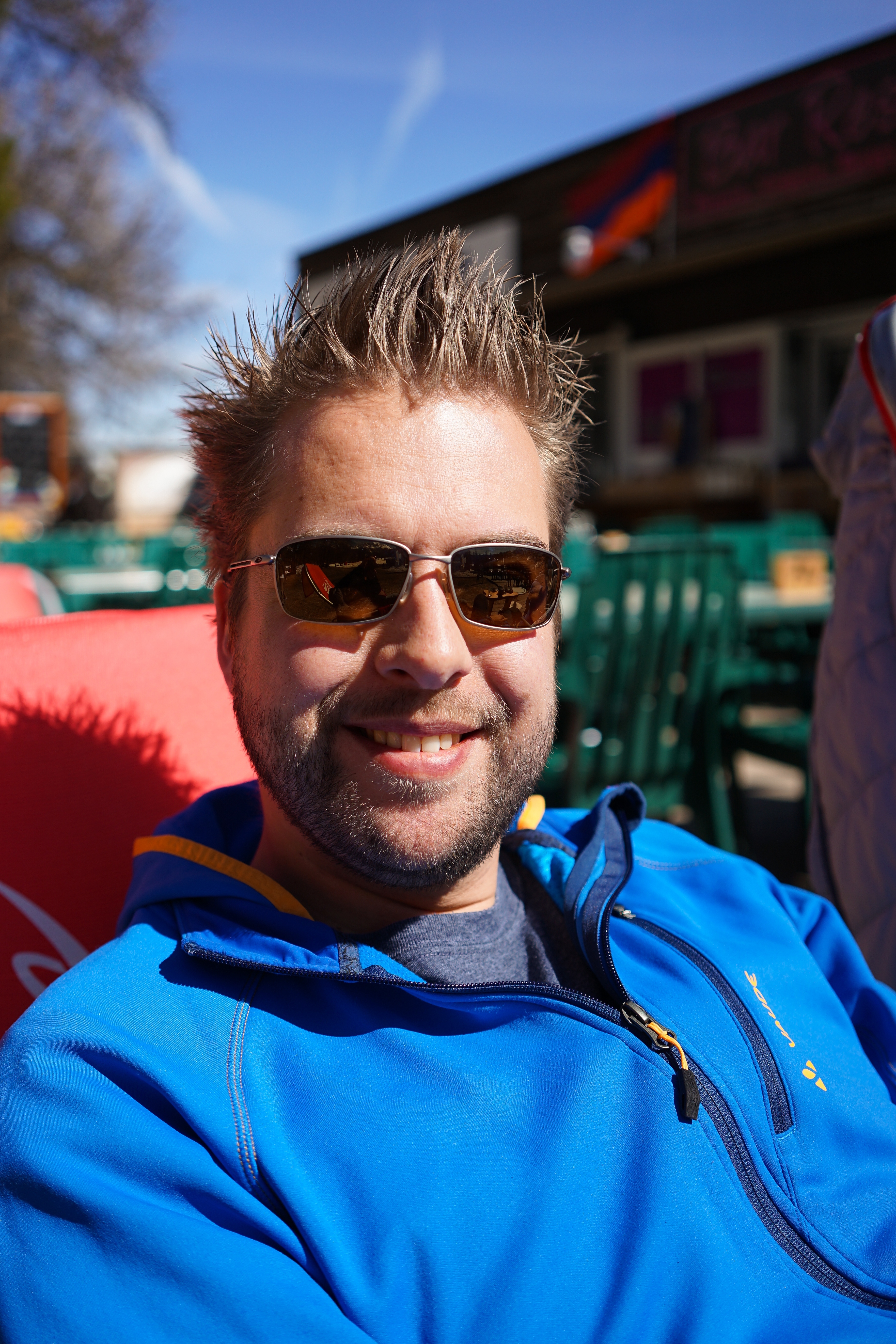 A man wearing sunglasses and a blue jacket smiles while sitting outdoors in the sunlight.