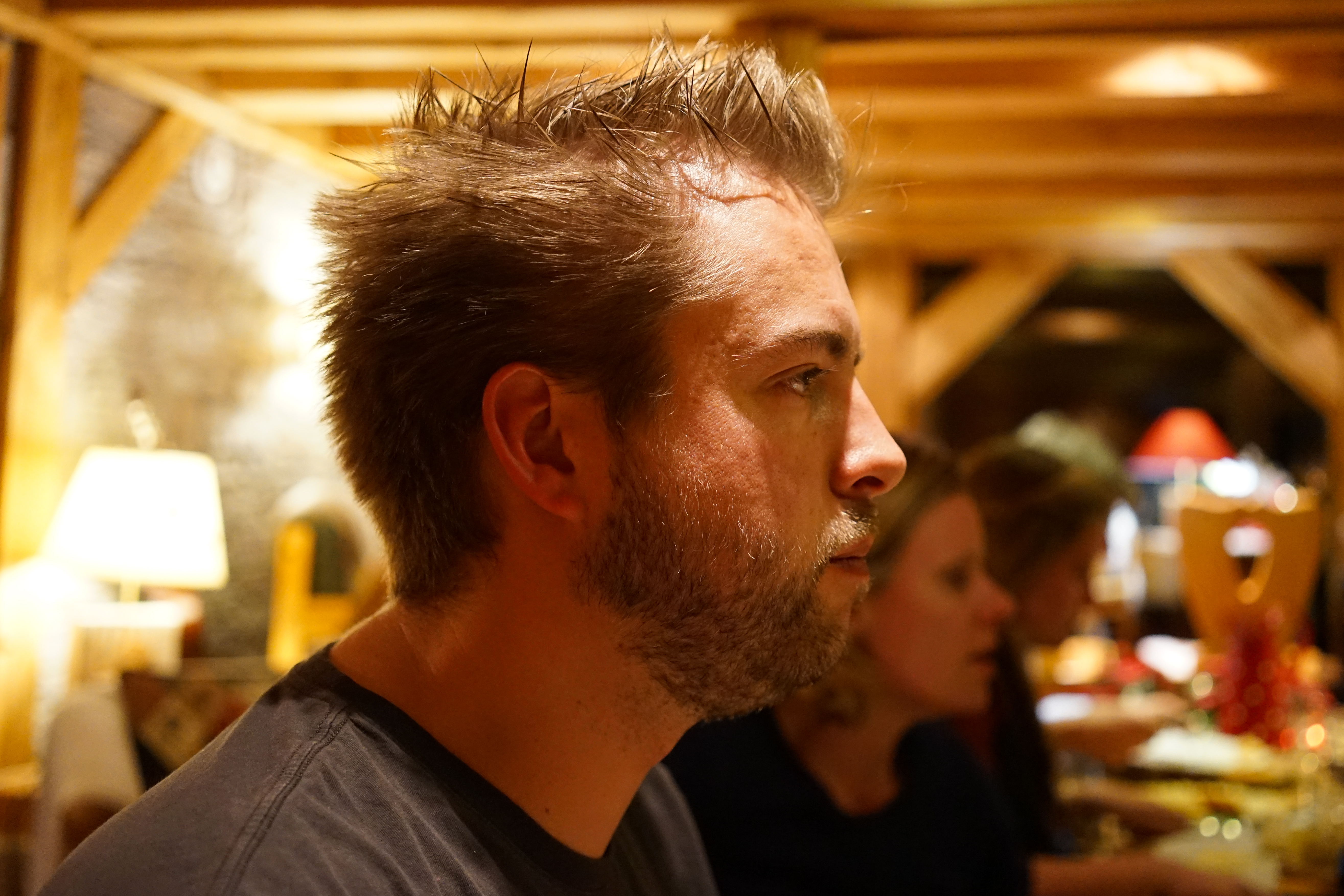 A man with spiky hair and a beard sits in a warmly lit restaurant, looking to the side.