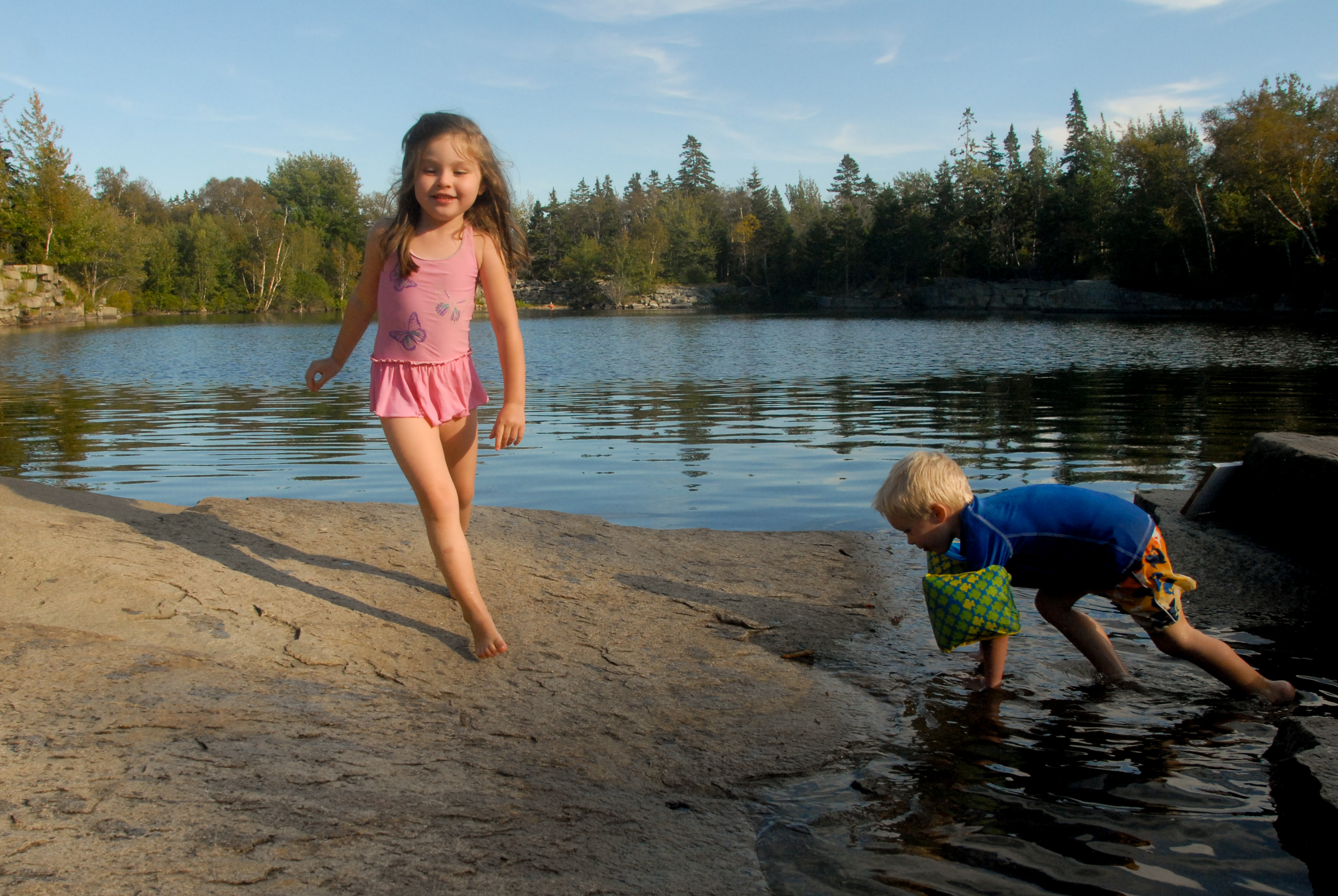 A young girl walks on a rocky surface while a boy crouches near the water, touching it with his hand.