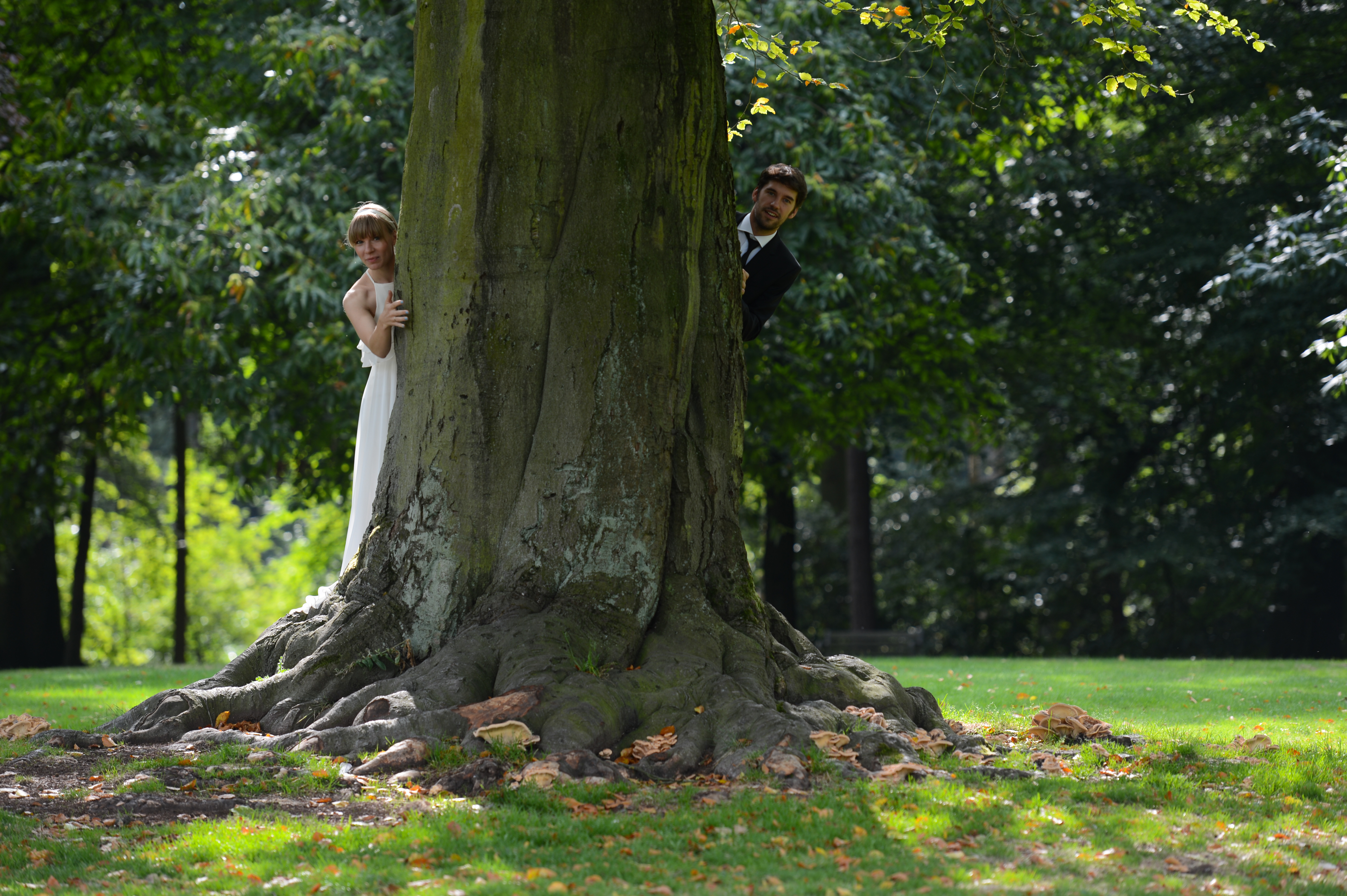 A bride and groom playfully peek out from behind a large tree in a green park.