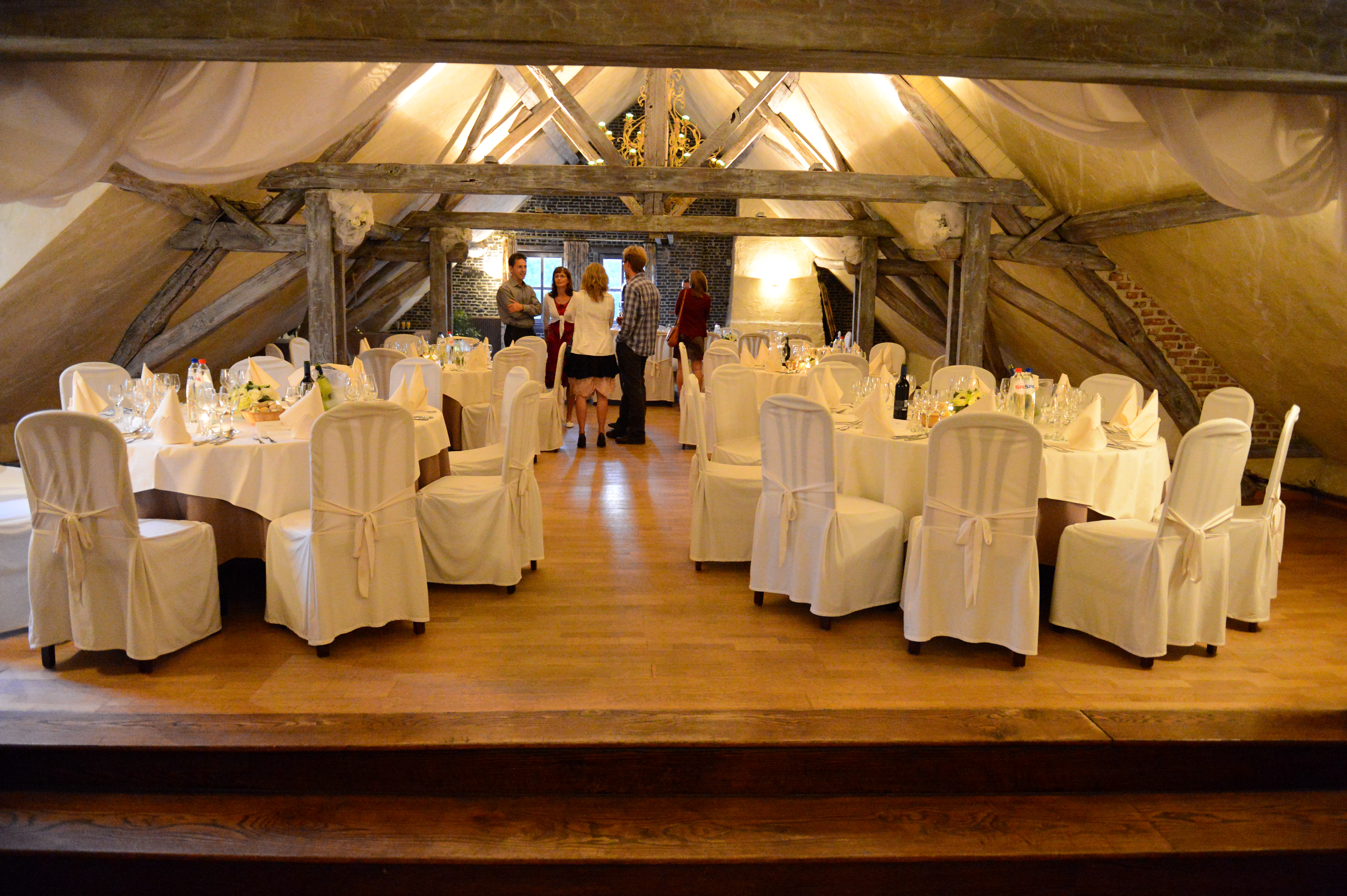 A decorated wedding reception hall with round tables and white chairs, as a small group stands and talks.