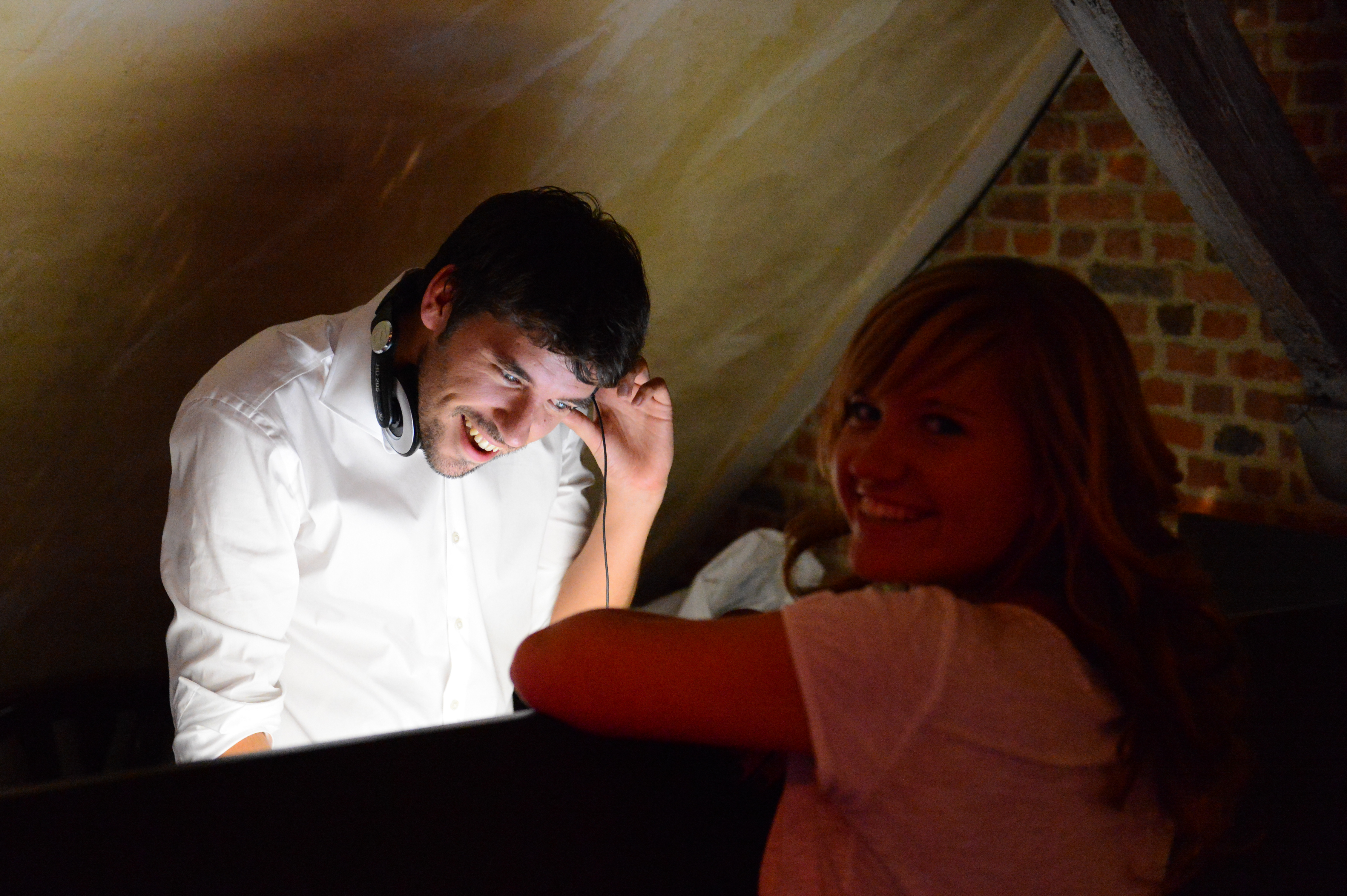 A smiling woman leans on a booth while a DJ in a white shirt adjusts his headphones and laughs.
