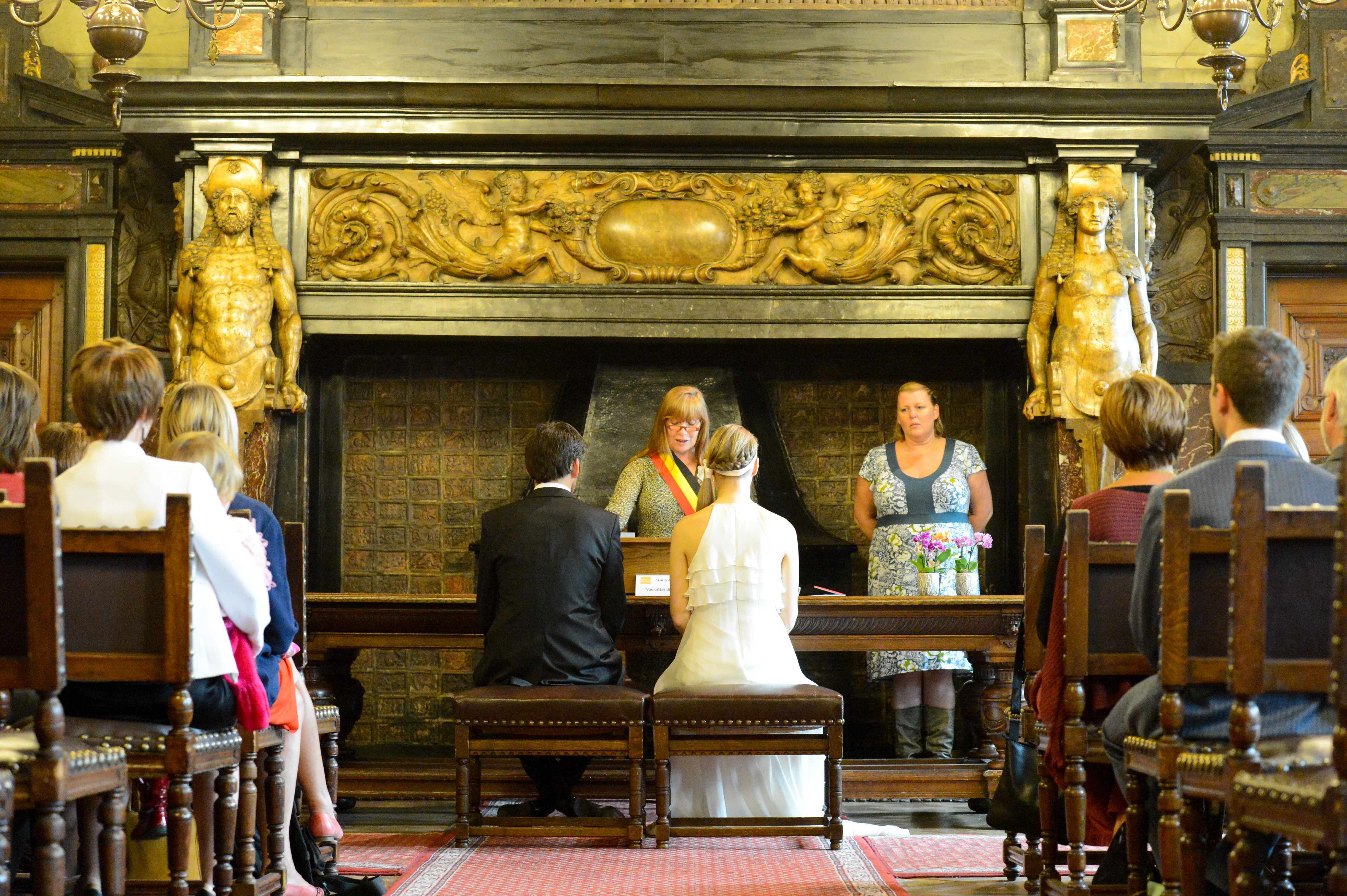 A bride and groom sit before an officiant during their wedding ceremony, surrounded by seated guests in a historic room.
