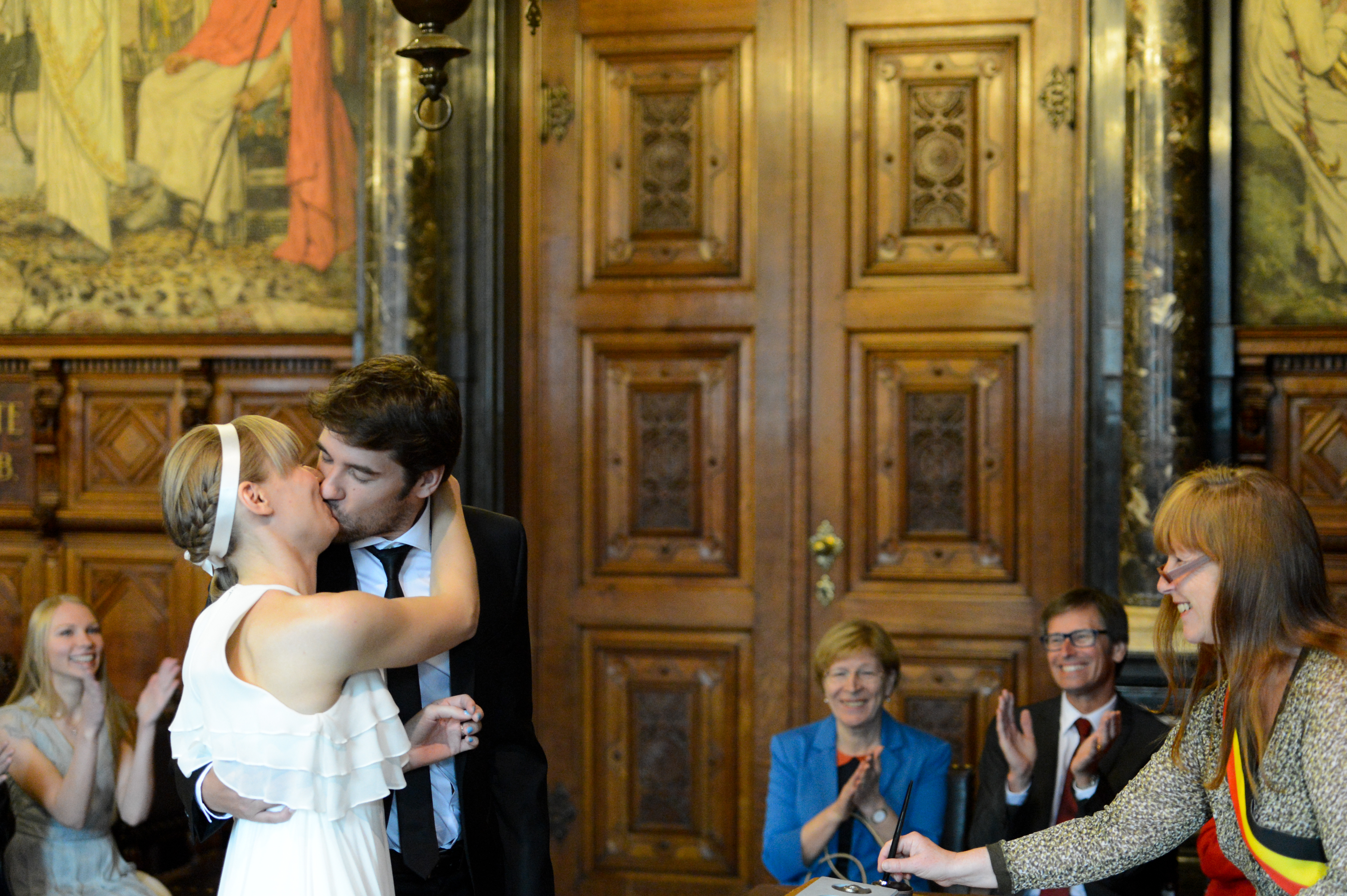 A newlywed couple shares a kiss while guests applaud in a formal wedding ceremony setting.