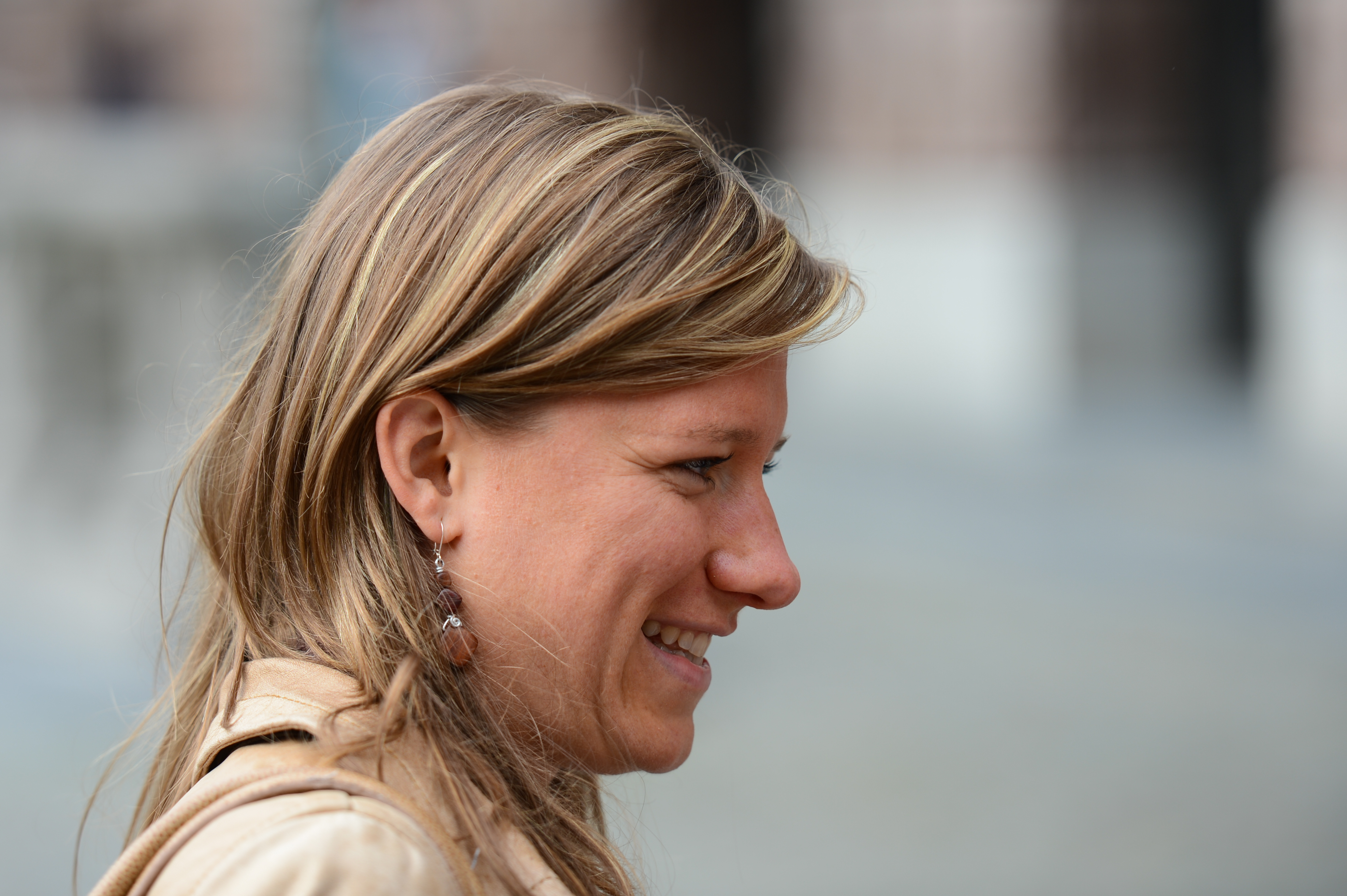 A woman with long blonde hair smiles while looking to the side, wearing a beige jacket and dangling earrings.