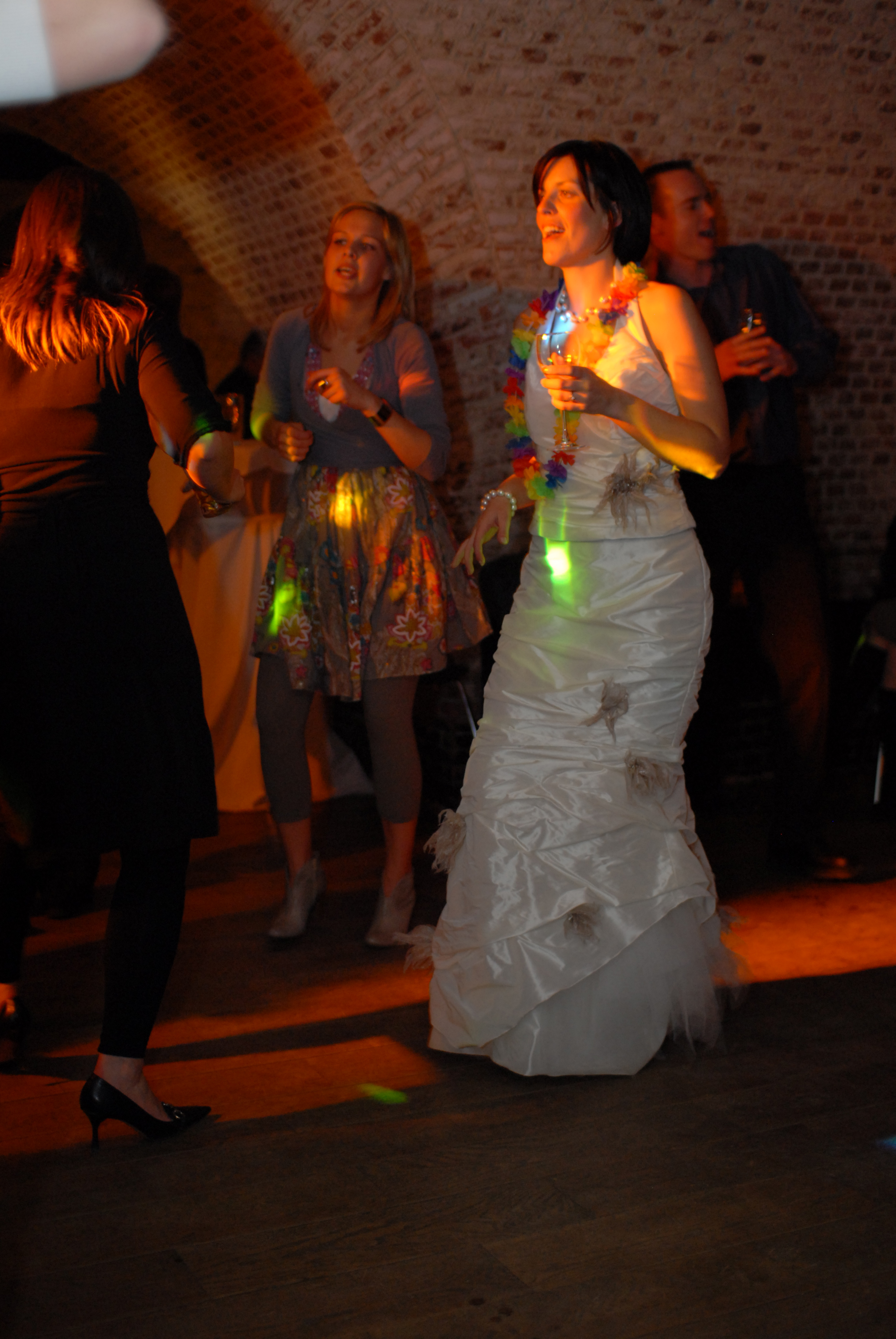 The bride in a white wedding dress dances with guests, holding a drink and wearing a colorful lei.
