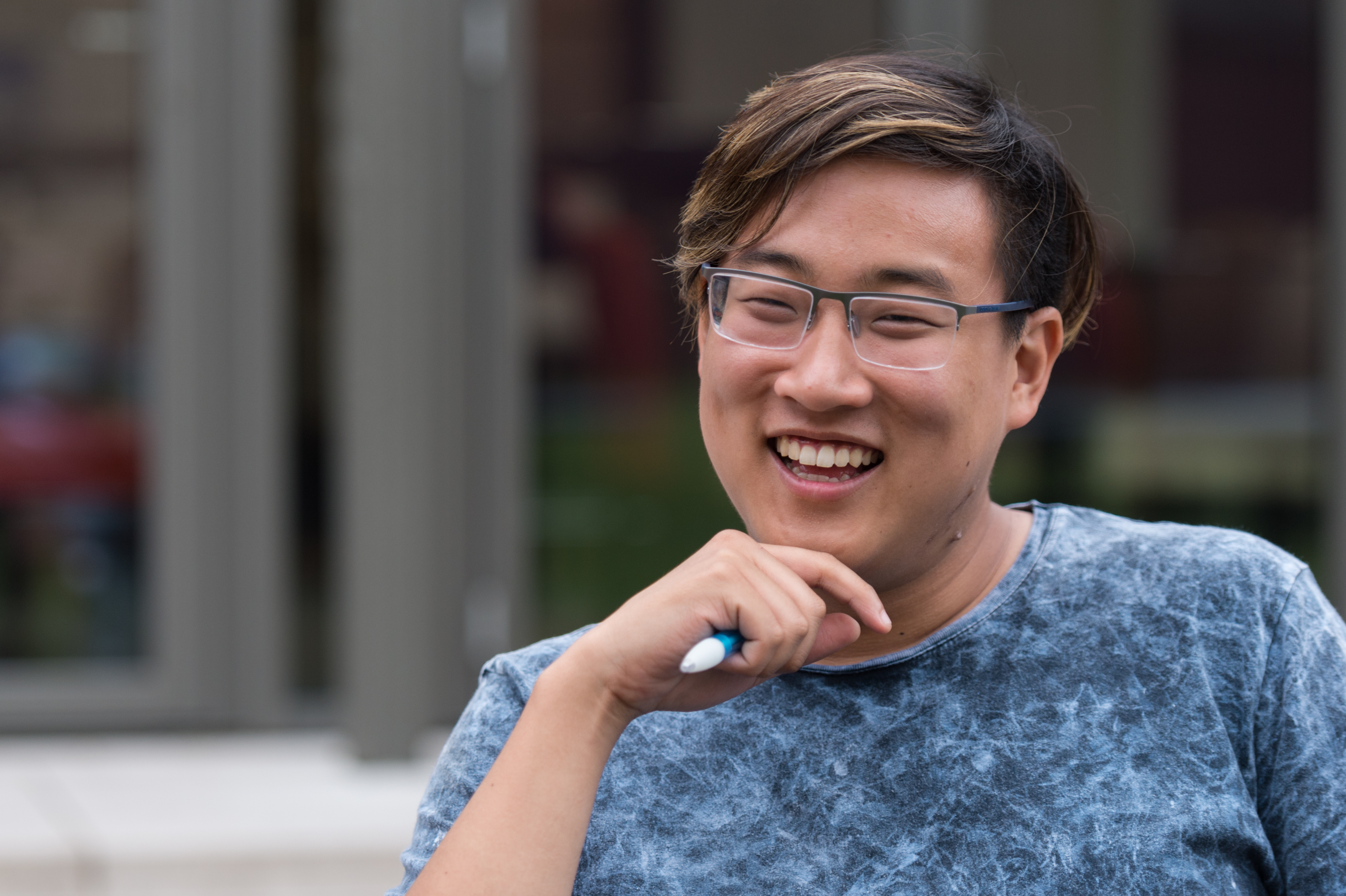 A smiling person wearing glasses and a blue shirt holds a pen while sitting outdoors.