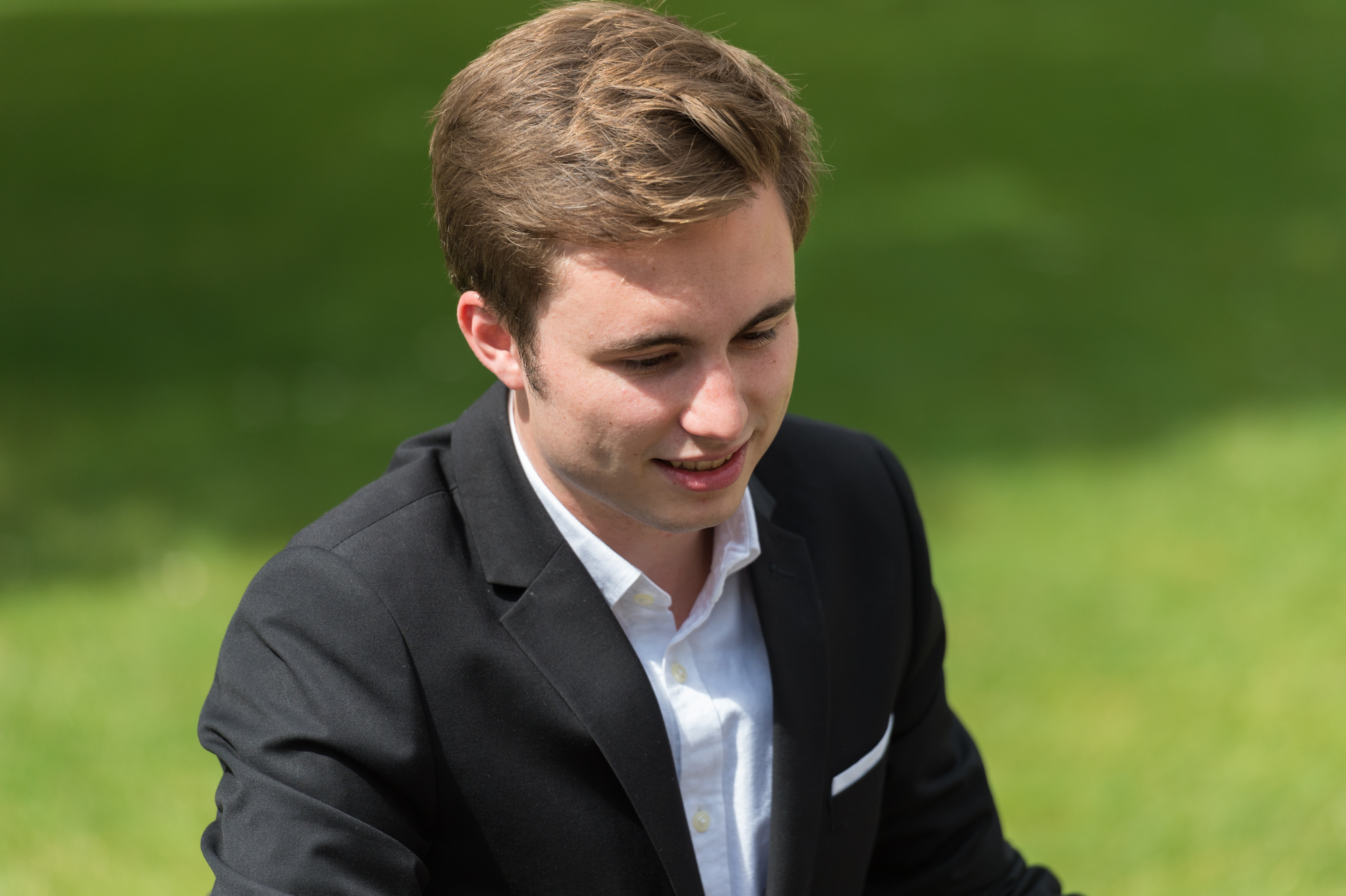 A man in a black suit and white shirt smiles while looking down at something outdoors.