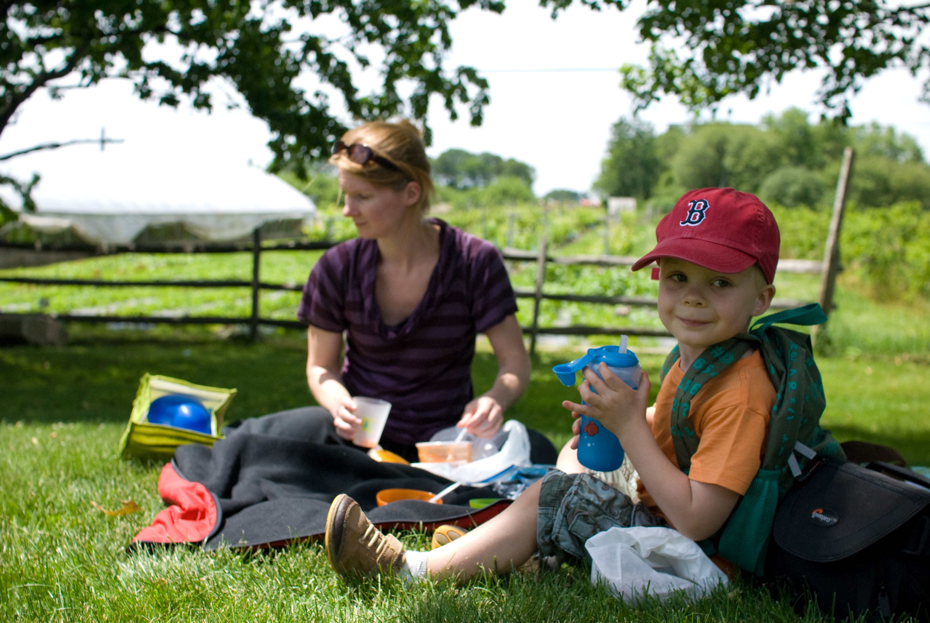 A child wearing a red cap sits on the grass, holding a drink, while an adult prepares food nearby.